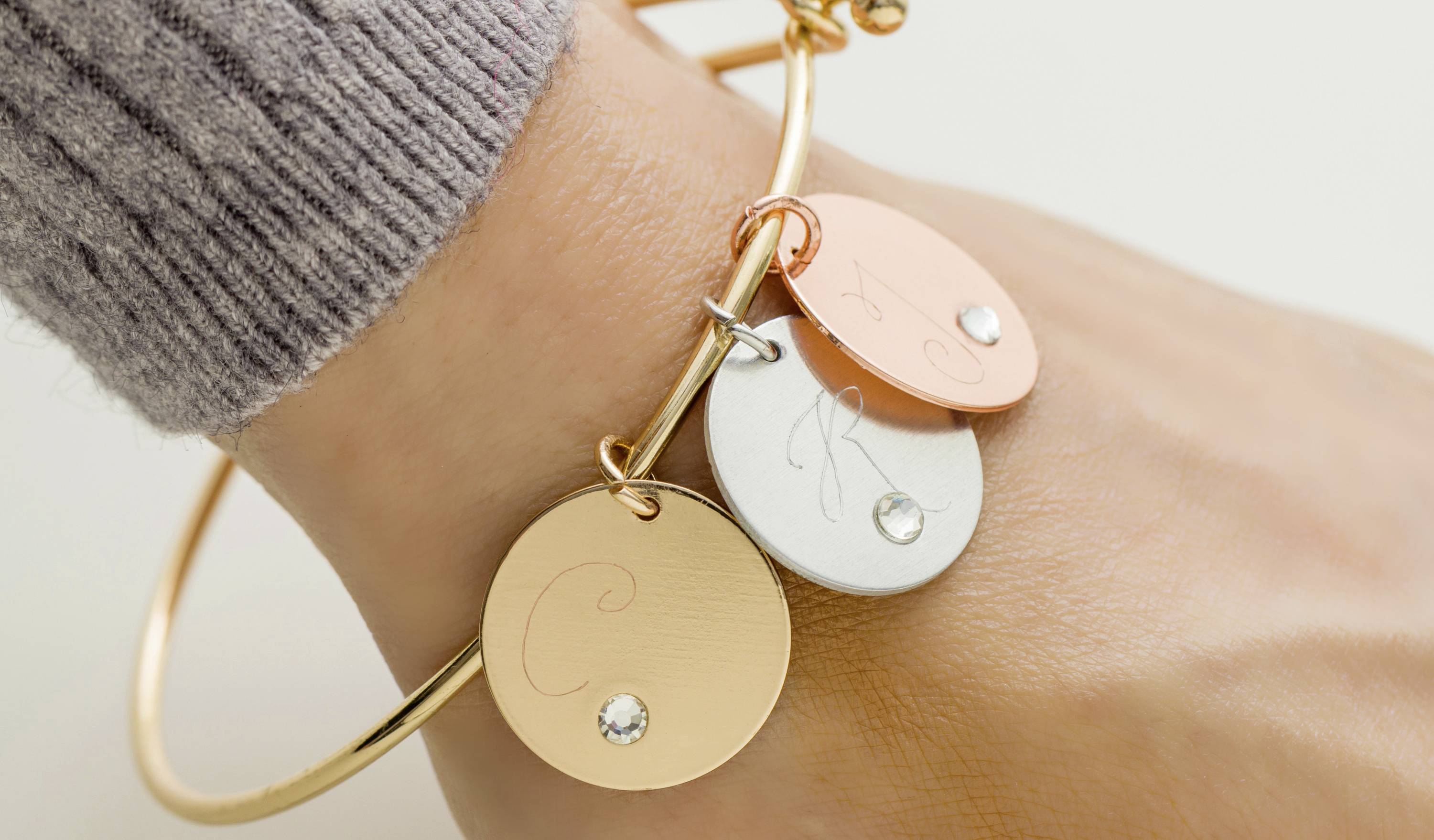 Armband mit drei runden Anhängern in Gold, Silber und Kupfer, jeweils mit gravierten Buchstaben und kleinen Diamanten verziert.