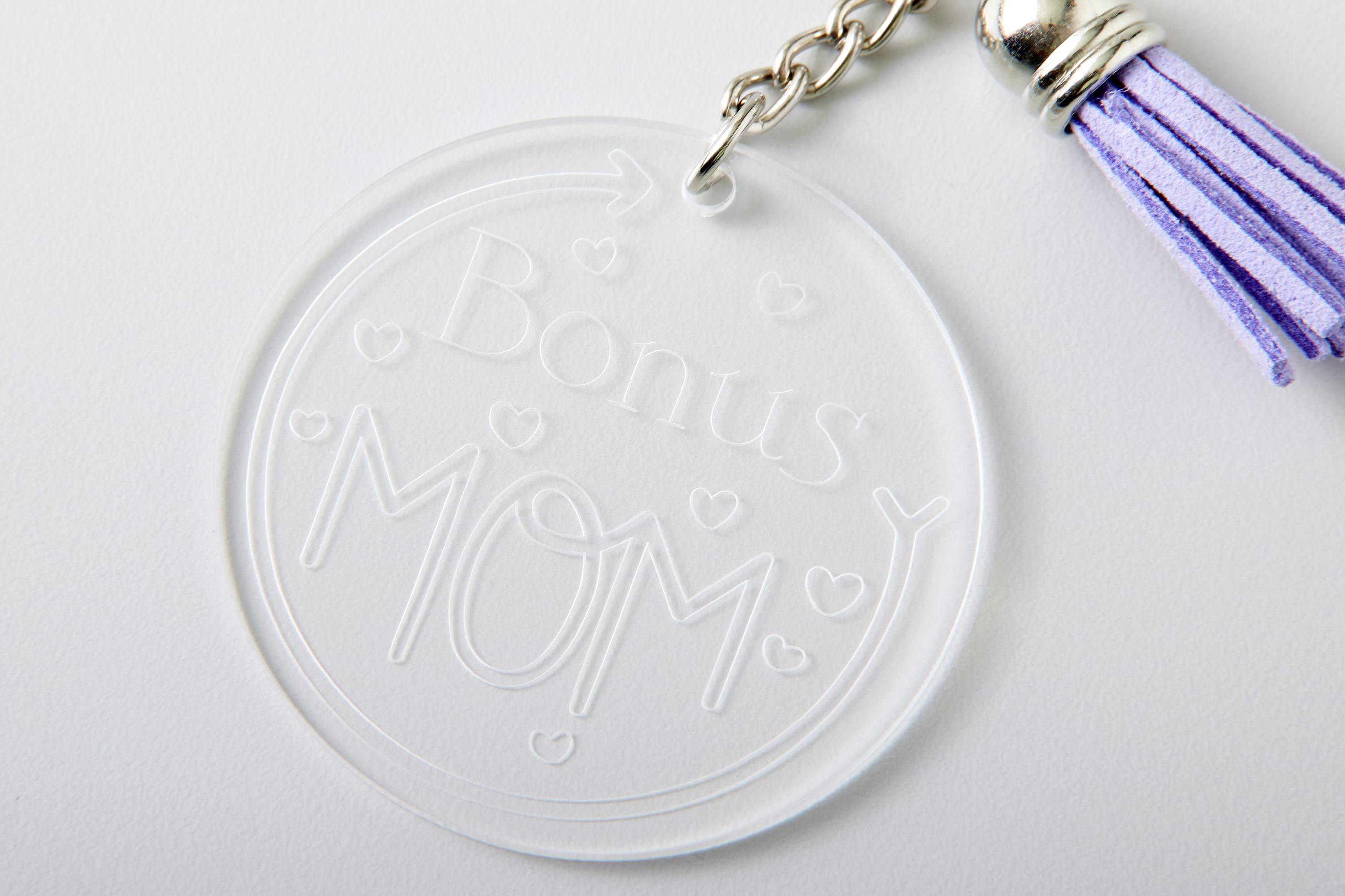 Schlüsselanhänger mit rundem, transparentem Anhänger, darauf steht 'Bonus Mom' in weißen Buchstaben, verziert mit kleinen Herzen, lila Quaste.