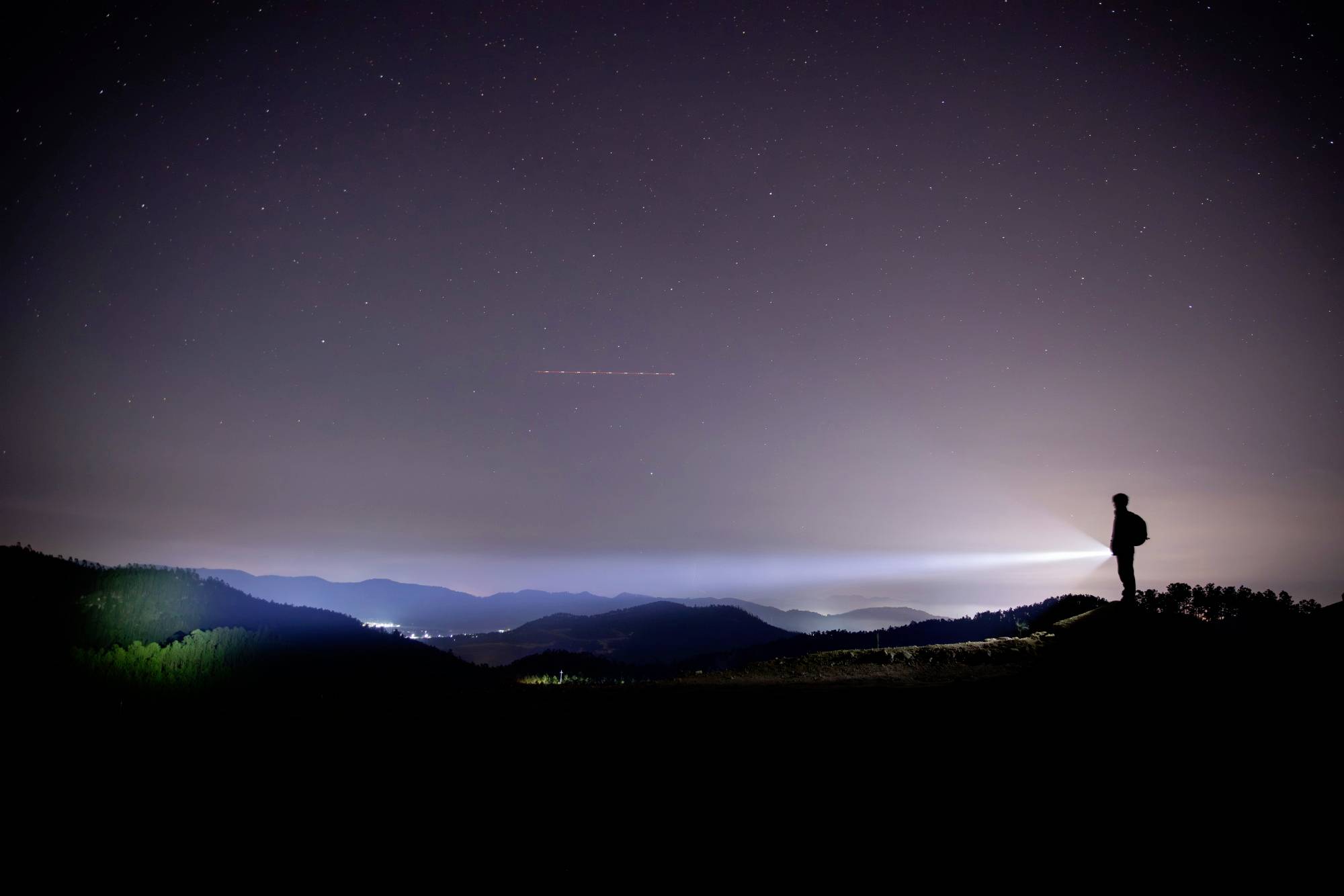 Eine Person steht auf einem Hügel in der Nacht, blickt in die Ferne und hält eine Taschenlampe, die einen Lichtstrahl wirft.