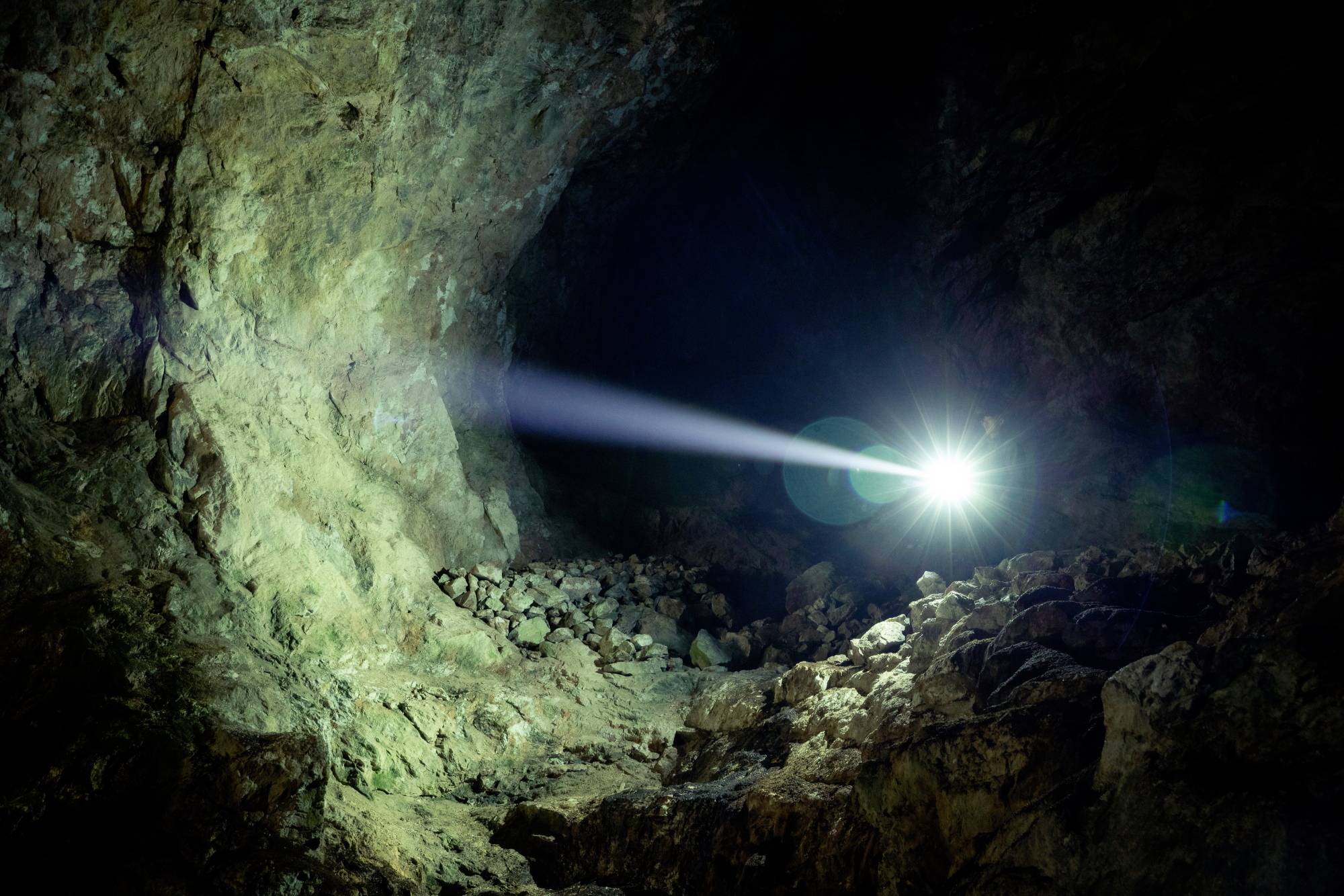 Ein heller Lichtstrahl leuchtet in eine dunkle Höhle, die steinige Wände und einen unebenen Boden zeigt.
