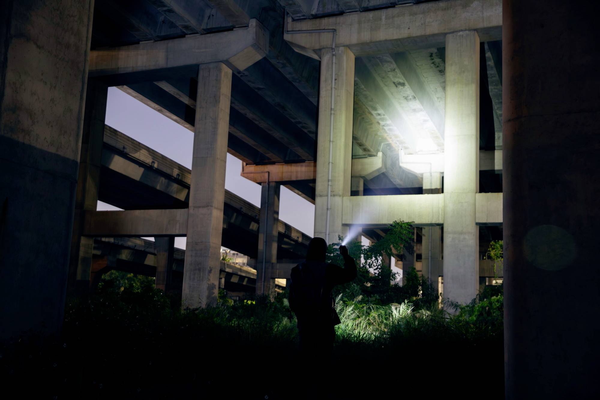 Eine Person steht nachts unter einer Autobahnbrücke und leuchtet mit einer Taschenlampe auf die Betonpfeiler.