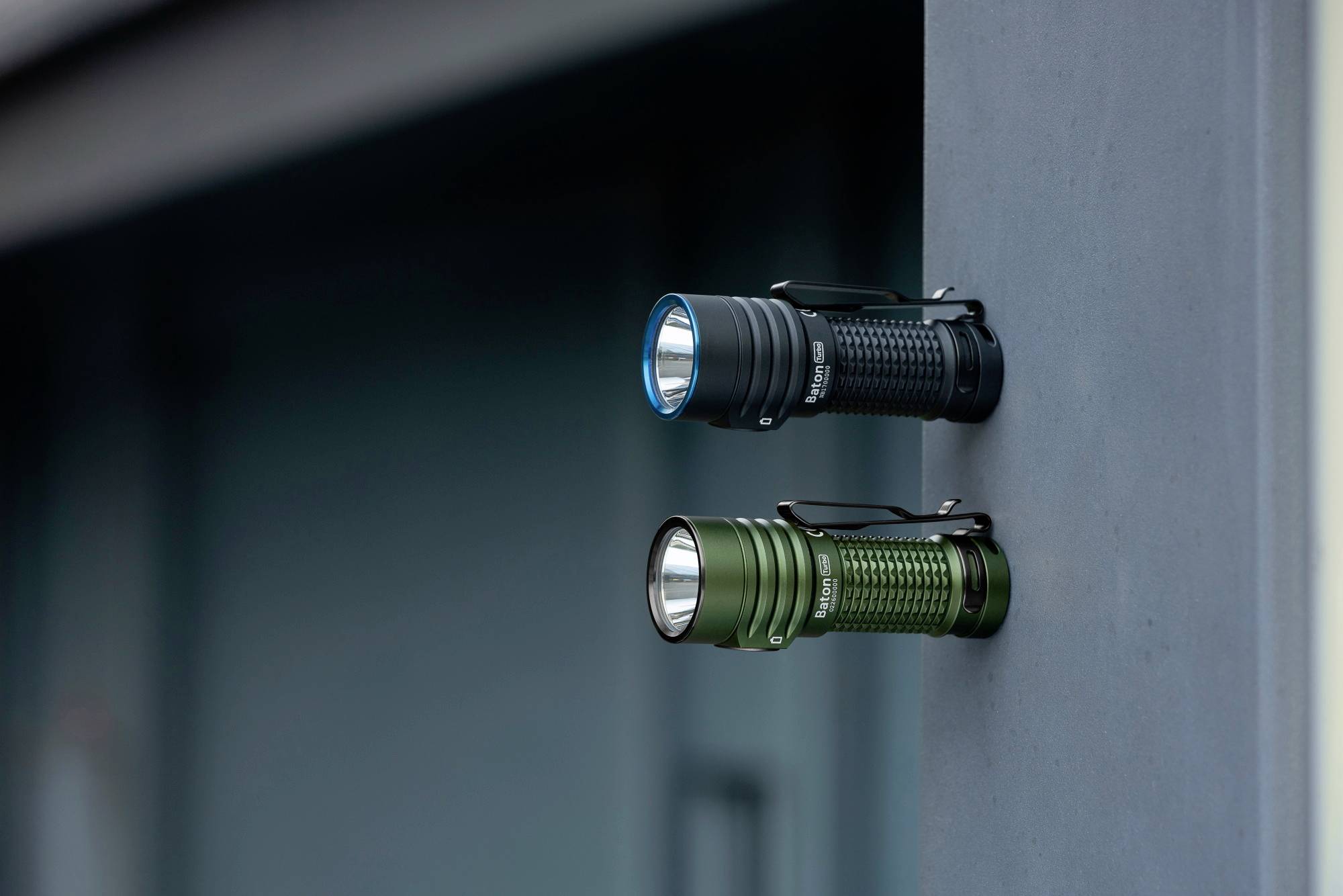 Zwei Taschenlampen, eine schwarz und eine grün, horizontal an einer grauen Metallfläche befestigt; beide mit strukturiertem Griff.
