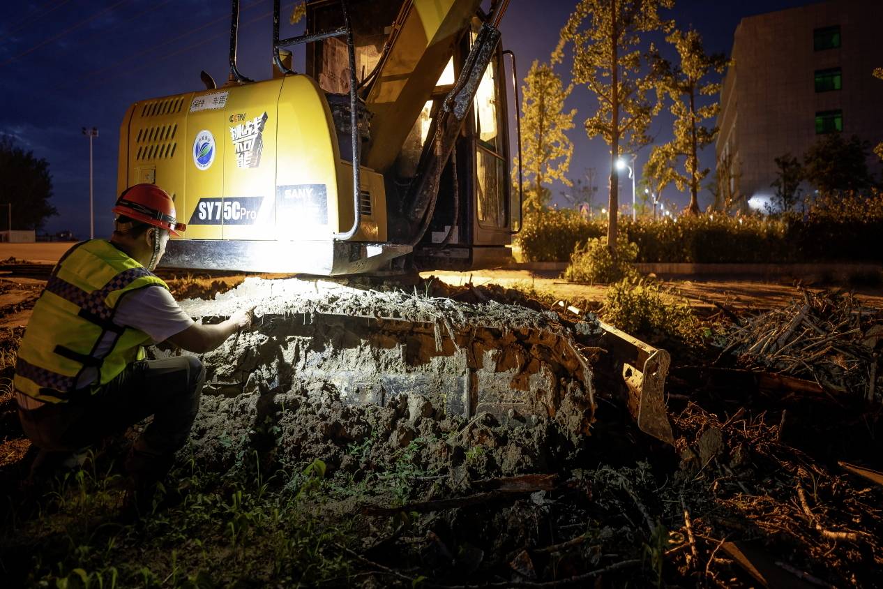 Eine Person in Arbeitskleidung überprüft nachts den unteren Teil eines gelben Baufahrzeugs, beleuchtet von Scheinwerfern, auf einer Baustelle.