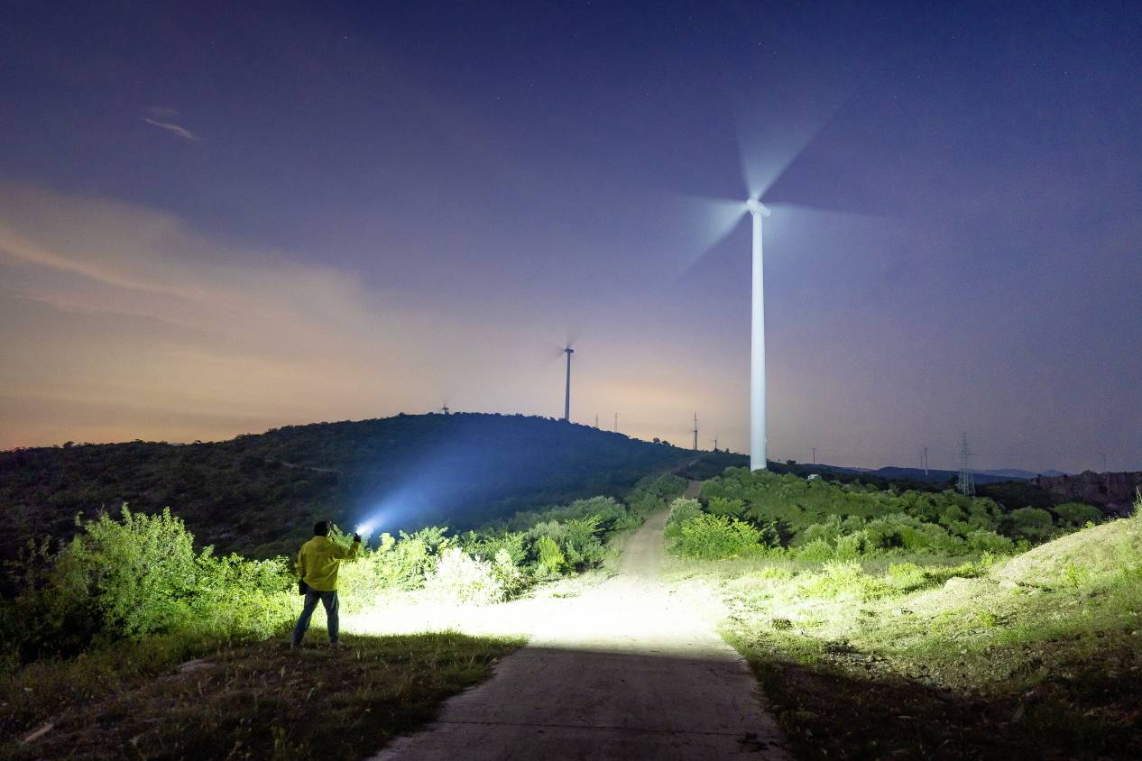 Eine Person mit Taschenlampe steht bei Dämmerung vor einem beleuchteten Windrad auf einem Hügel, umgeben von weiteren Windrädern.