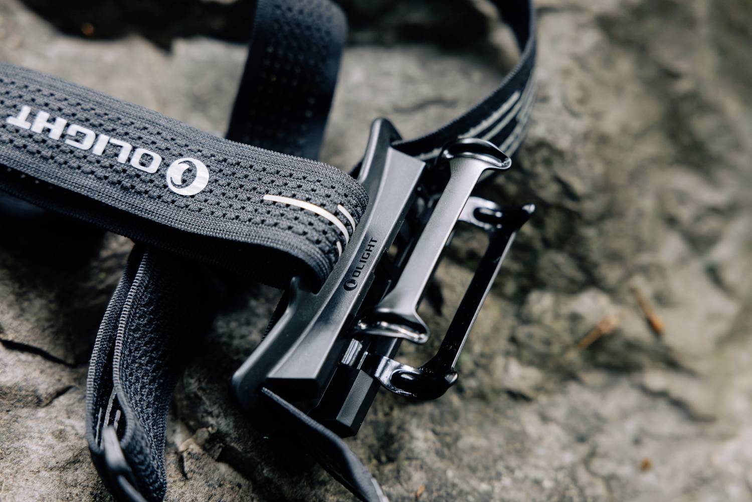 Eine schwarze Gürtelschnalle mit der Aufschrift 'Olight' liegt auf einem grauen, unebenen Stein.