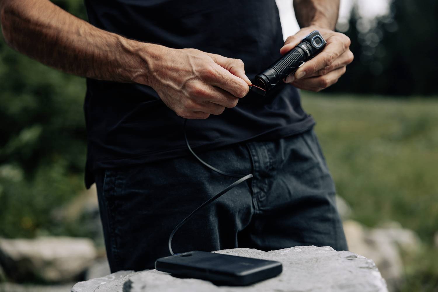 Eine Person hält eine Taschenlampe und verbindet sie mit einem schwarzen tragbaren Ladegerät, während sie draußen in der Natur steht.