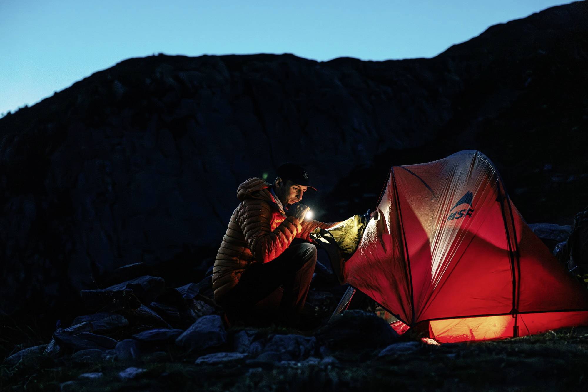 Person bei Nacht mit Taschenlampe im Gebirge neben einem beleuchteten Zelt.