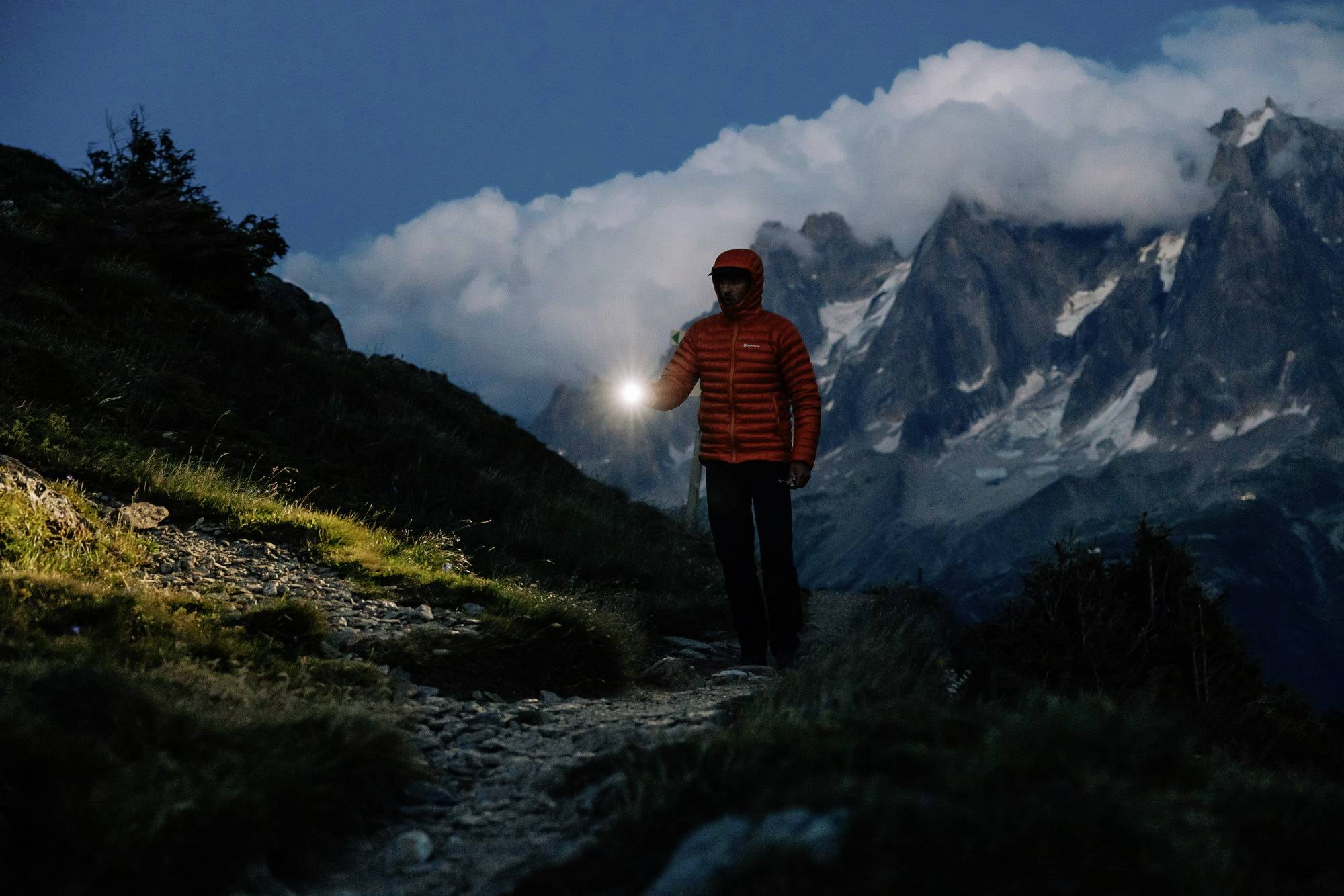 Eine Person wandert bei Dämmerung mit Taschenlampe auf einem Bergpfad, im Hintergrund sind wolkenverhangene Gipfel zu sehen.