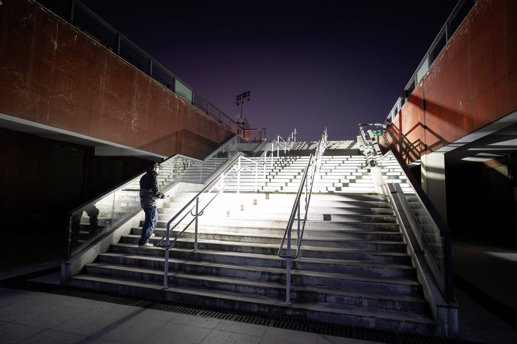 Eine Person steigt eine beleuchtete Treppe hinauf, während eine andere Person am Geländer ruht. Es ist Nacht, und die Umgebung ist ruhig.