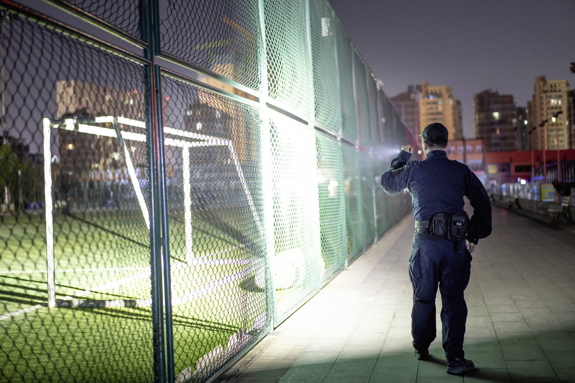 Ein Sicherheitsbeamter patrouilliert nachts neben einem eingezäunten Fußballfeld in einer Stadtumgebung.