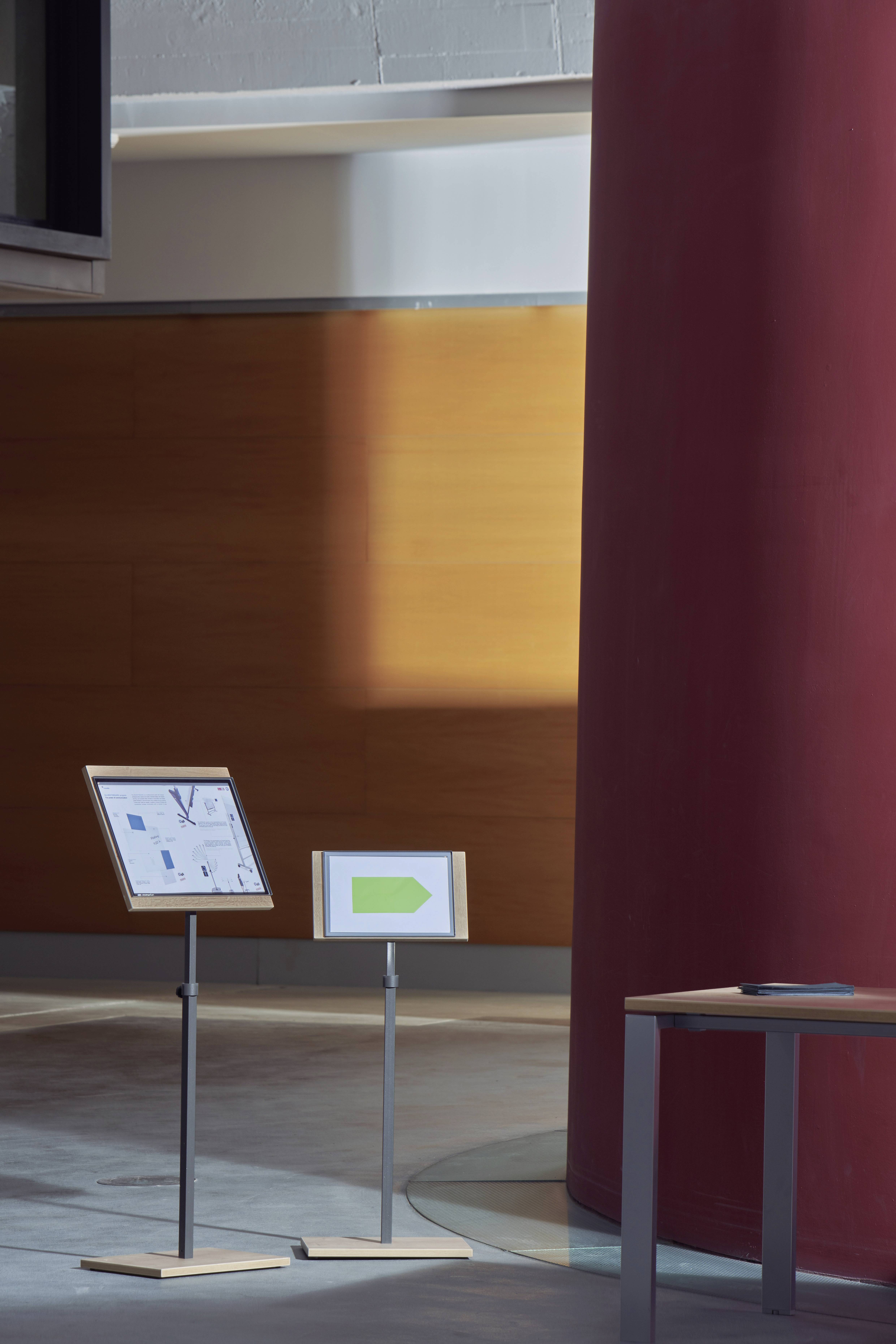 Zwei Tafeln stehen in einer modernen Halle. Links zeigt ein kleiner Übersichtsplan, rechts ein Schild mit einem grünen Pfeil.