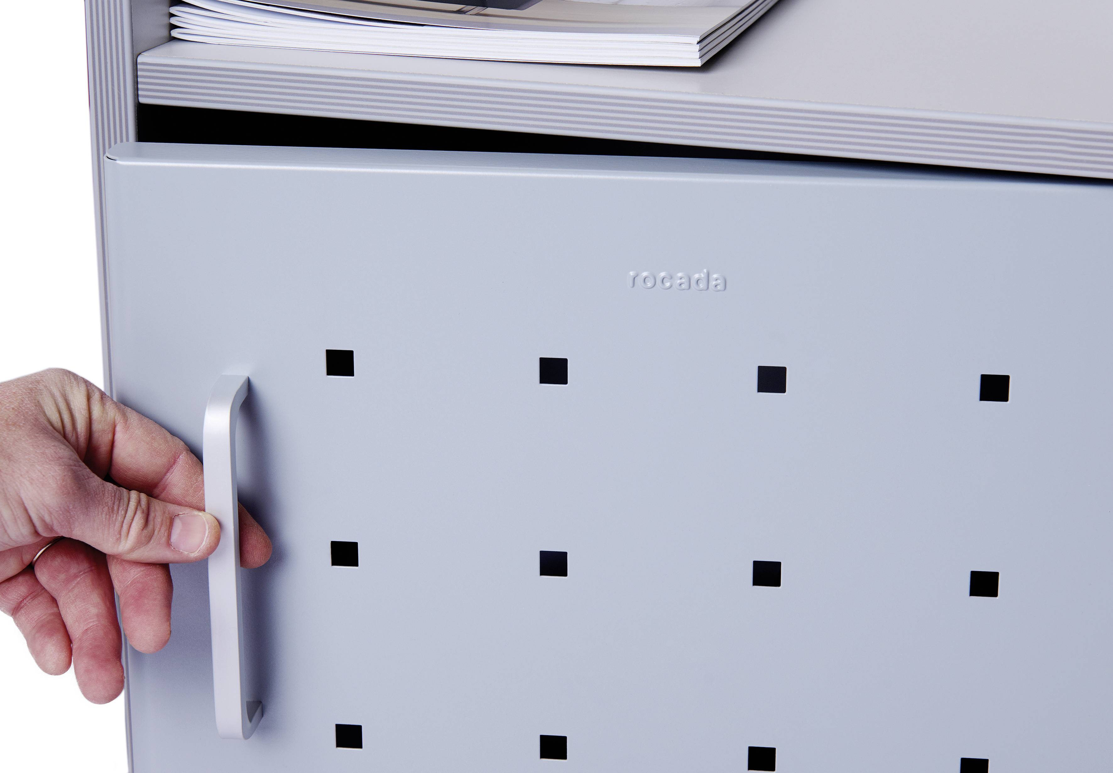 Hand zieht an einem quadratisch gelochten Türgriff eines grauen Metallschranks. Oben liegen Zeitschriften auf dem Schrank.