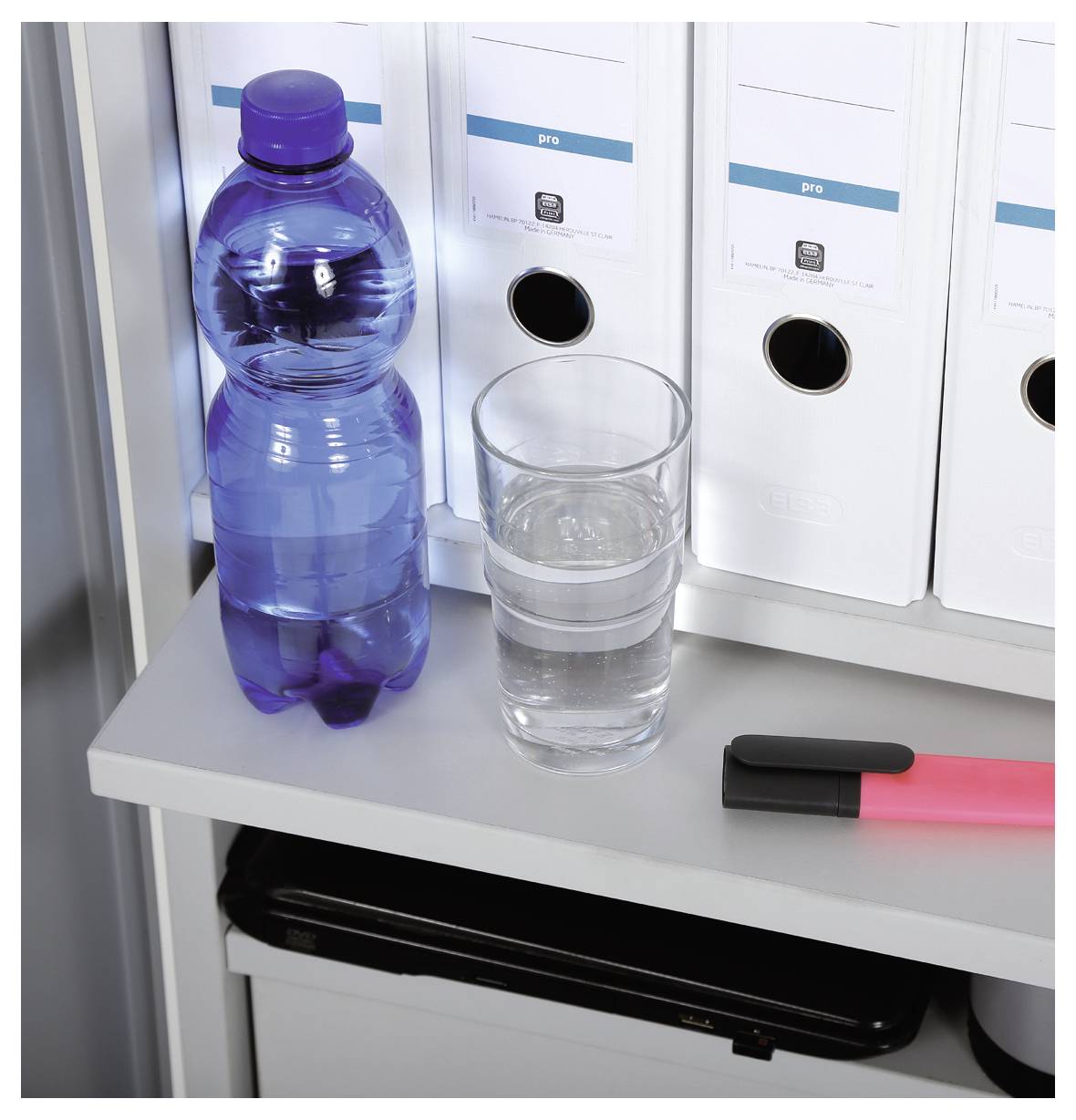 Auf einem Regal stehen eine vollgefüllte Wasserflasche, ein halbleeres Glas mit klarem Wasser, ein rosa Textmarker und weiße Ordner.