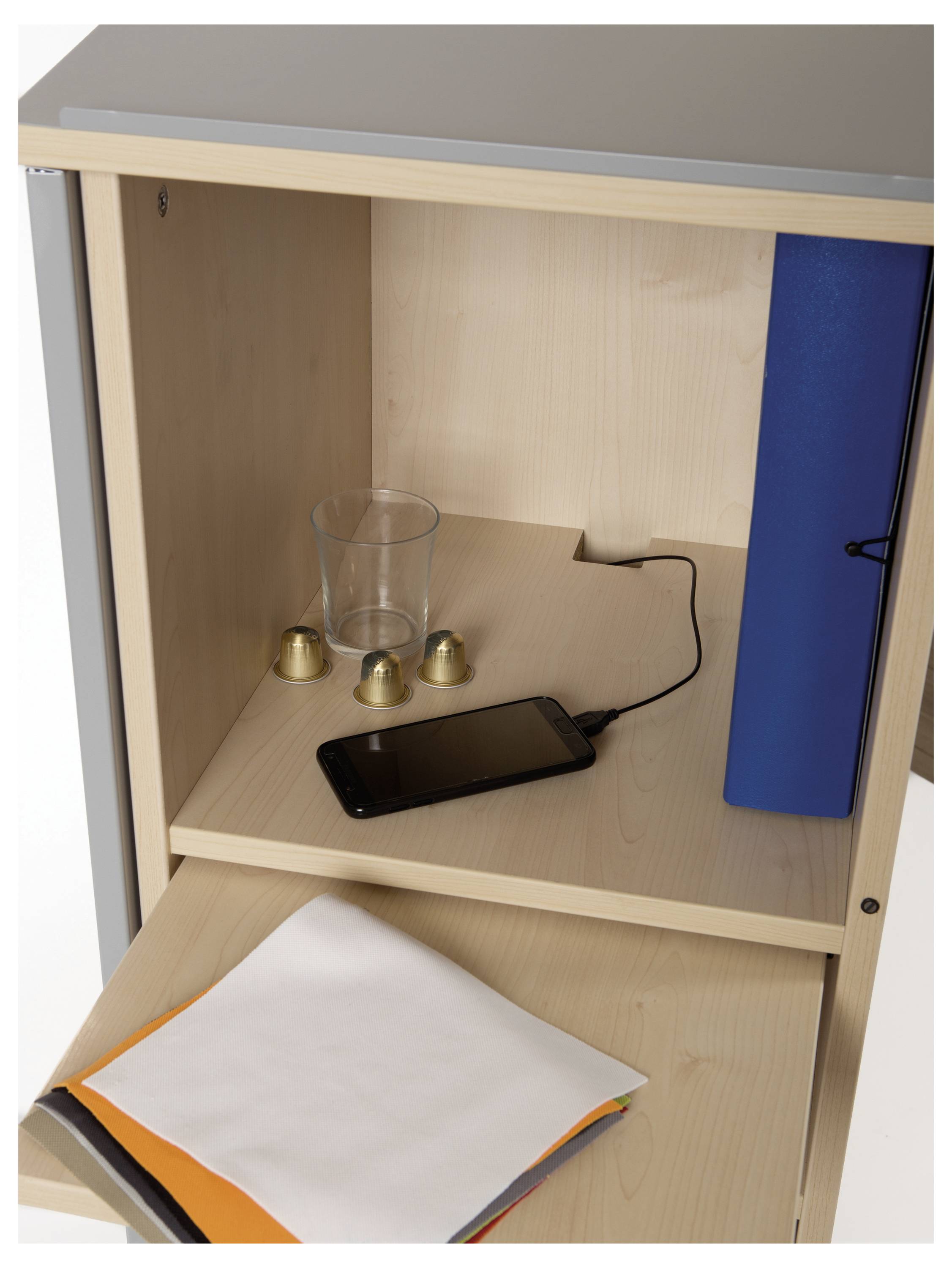 Ein offener Holzschrank mit einem Glas, drei kleinen Metallglocken, einem blauen Ordner und einem Smartphone, das aufgeladen wird.