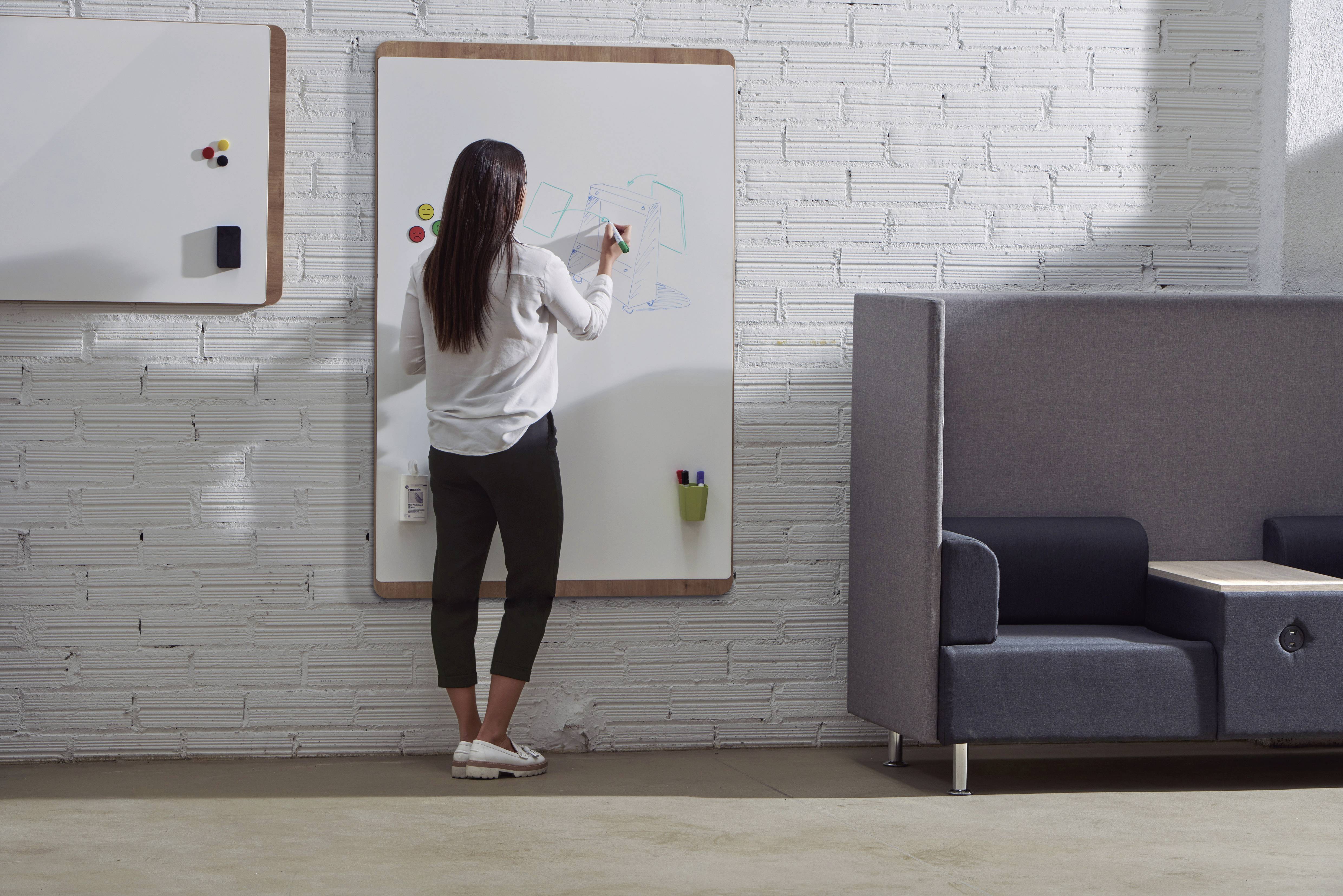 Eine Frau zeichnet auf einem Whiteboard in einem modernen Büro. Im Hintergrund ein Sofa und eine weiße Backsteinwand.