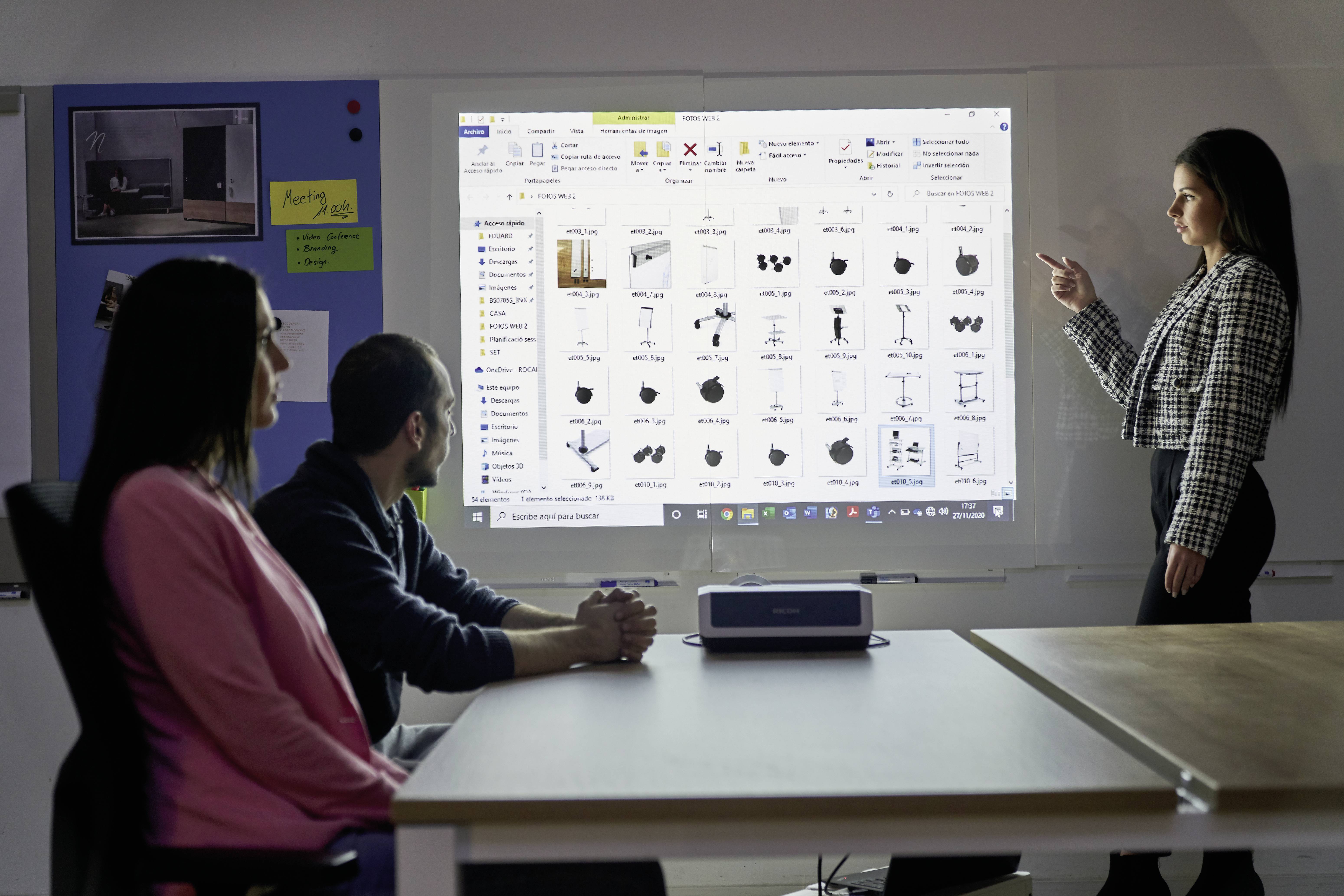 Eine Frau steht und präsentiert, während zwei Personen am Tisch sitzen und zuhören, mit Bildern auf der Leinwand im Hintergrund.