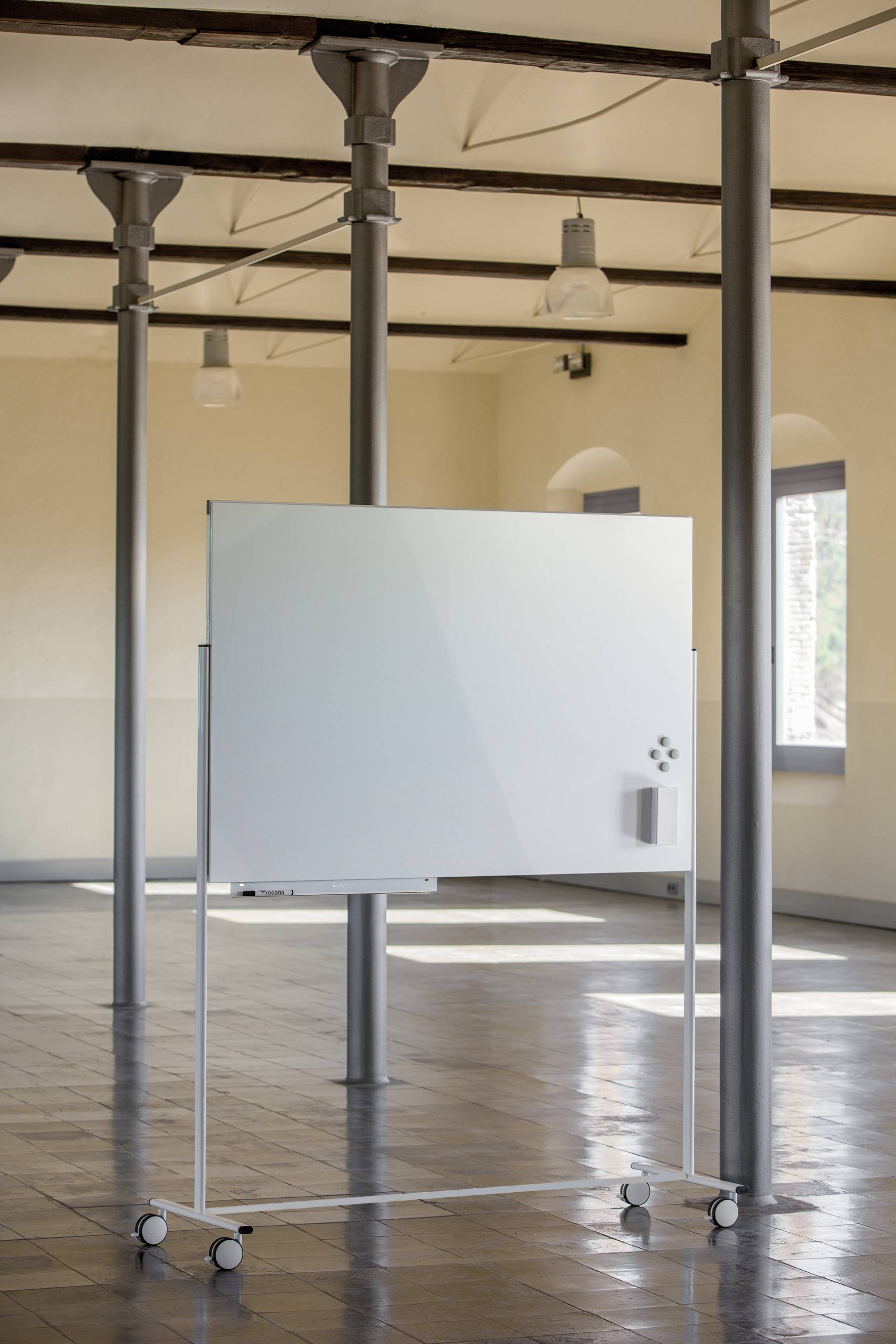 Mobiles Whiteboard auf Rollen steht in einem großen, hellen Raum mit Fliesenboden und Säulen. Ideal für Präsentationen oder Workshops.