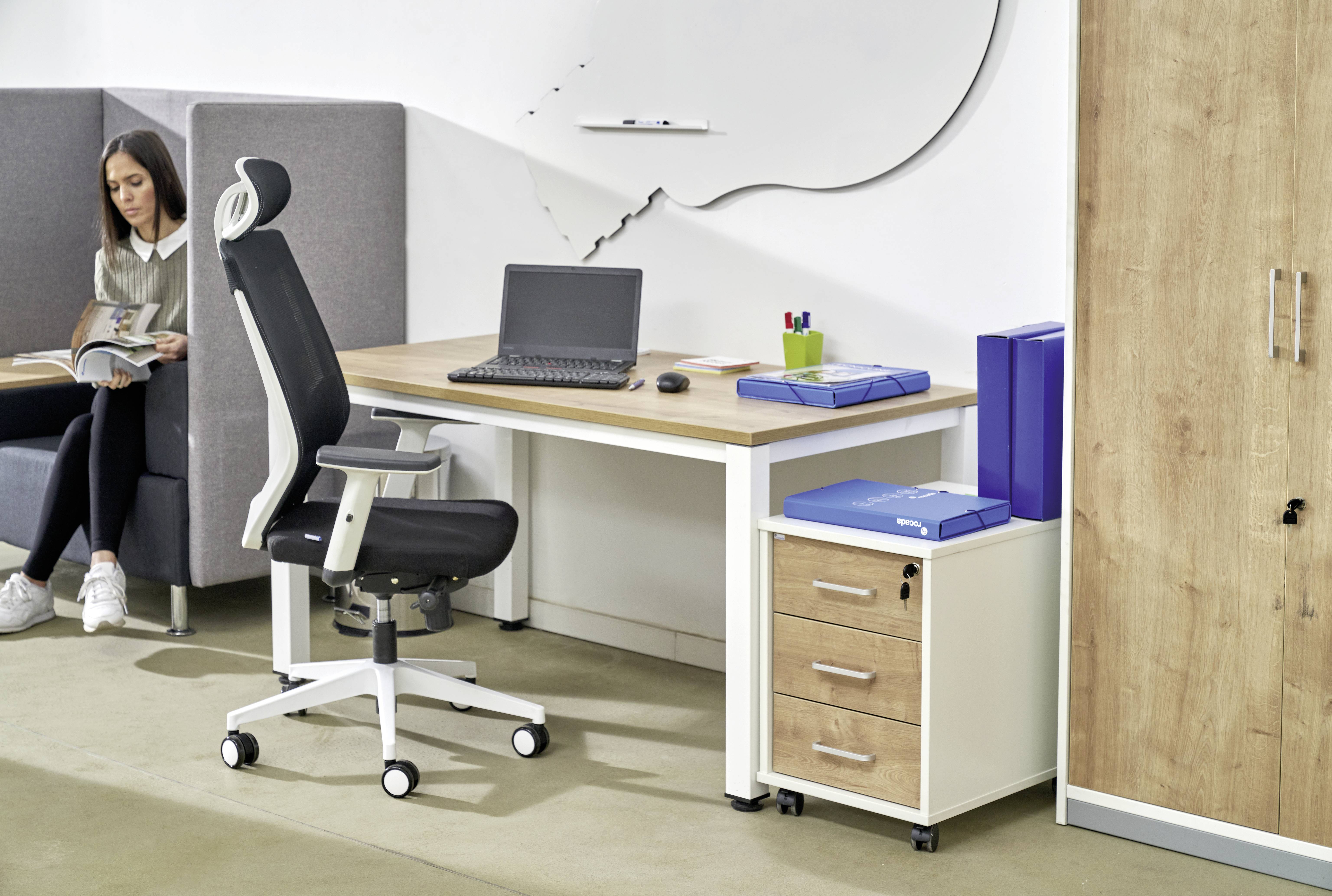 Ein modernes Büro mit einem Schreibtisch, auf dem ein Laptop steht. Daneben eine ergonomische Bürostuhl und ein Rollcontainer. Im Hintergrund sitzt eine Frau auf einem Sofa und liest ein Buch.