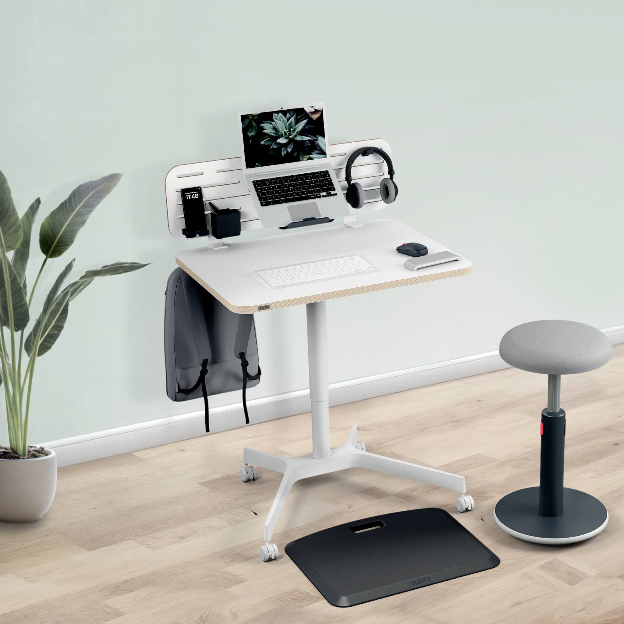 Ein moderner, höhenverstellbarer Schreibtisch mit Laptop, Kopfhörern und ergonomischem Hocker in einem hellen Raum mit Pflanzen.