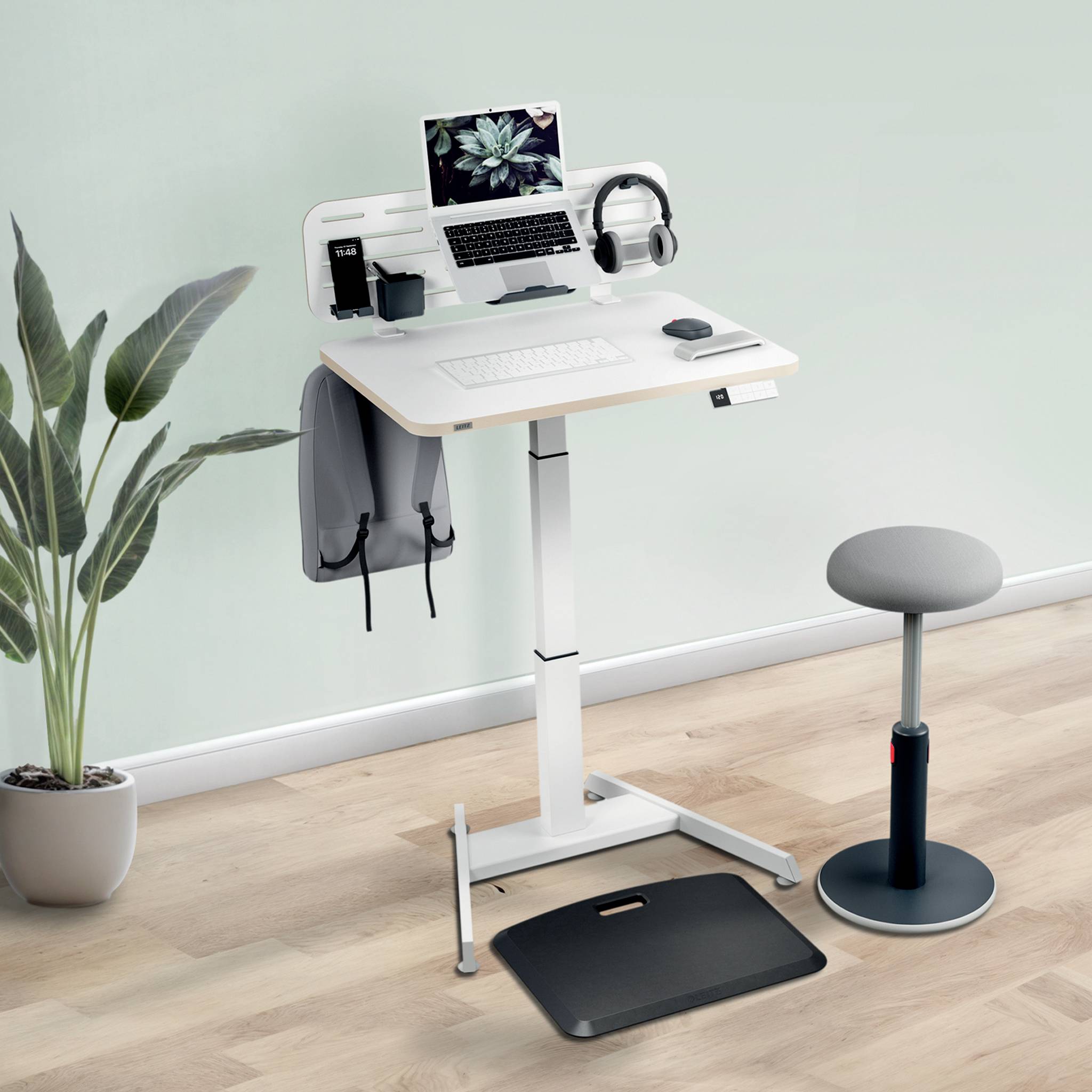 Ein höhenverstellbarer Schreibtisch mit einem Laptop, Kopfhörer und Schreibtischzubehör, umgeben von einer Pflanze und einem Hocker.