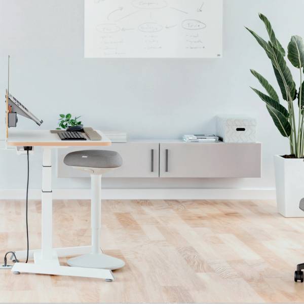 Ein höhenverstellbarer Schreibtisch mit Laptop, Stuhl und Pflanze. Im Hintergrund ein Whiteboard mit Diagramm und ein Regal.