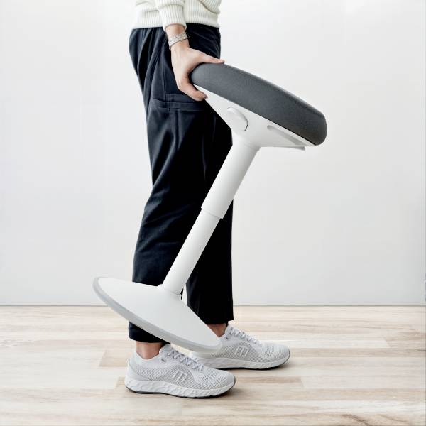 Person hält einen modernen ergonomischen Stuhl mit schräger Sitzfläche in einem hellen Raum. Der Stuhl hat ein minimalistisches Design.