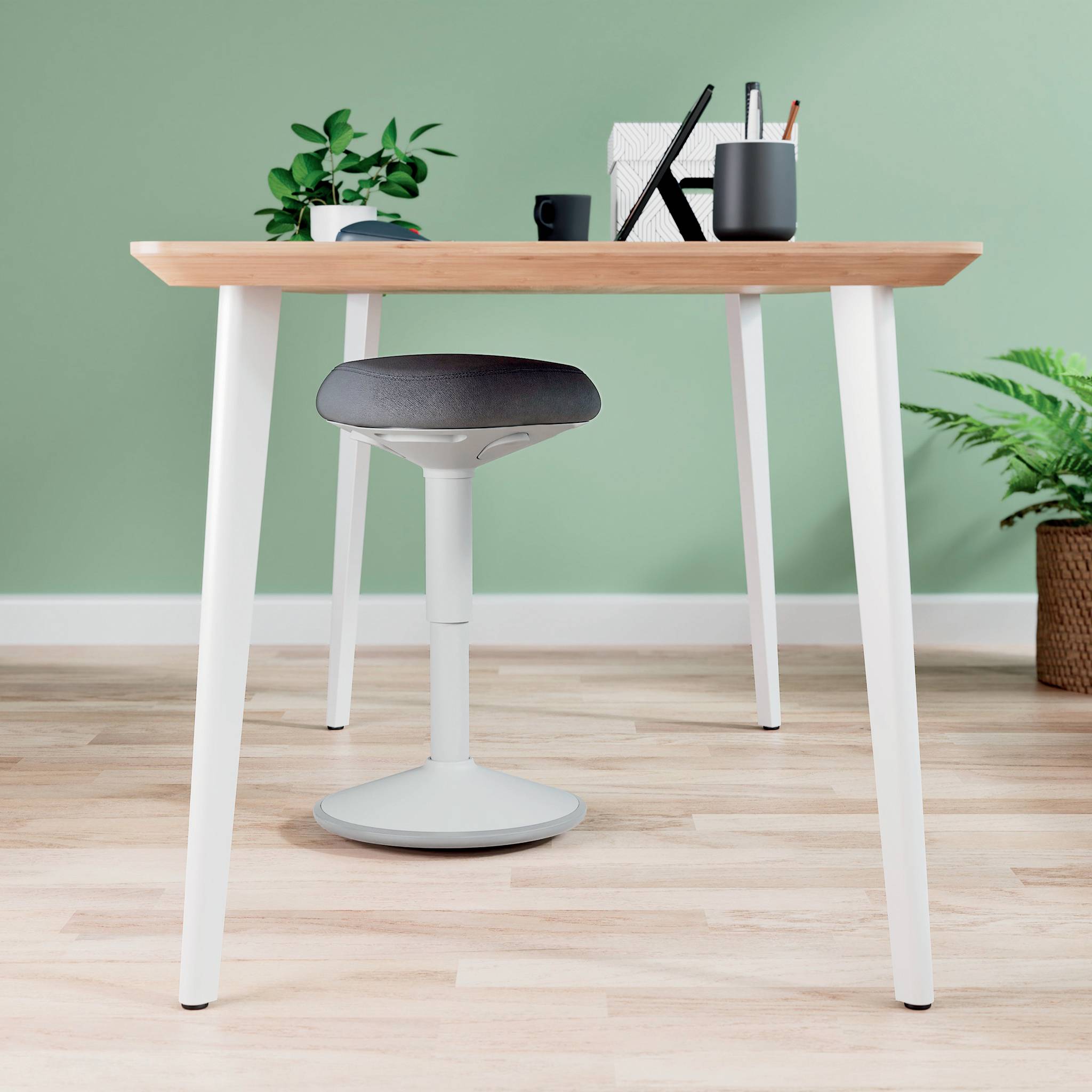 Schreibtisch mit Holzplatte, Büropflanzen, Büromaterialien und einem ergonomischen Hocker auf heller Holzbodenfläche vor grüner Wand.