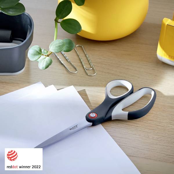 Eine Schere liegt auf einem Holztisch mit weißen Papierblättern und Büroklammern. Im Hintergrund sind Pflanze und gelbe Vase zu sehen.