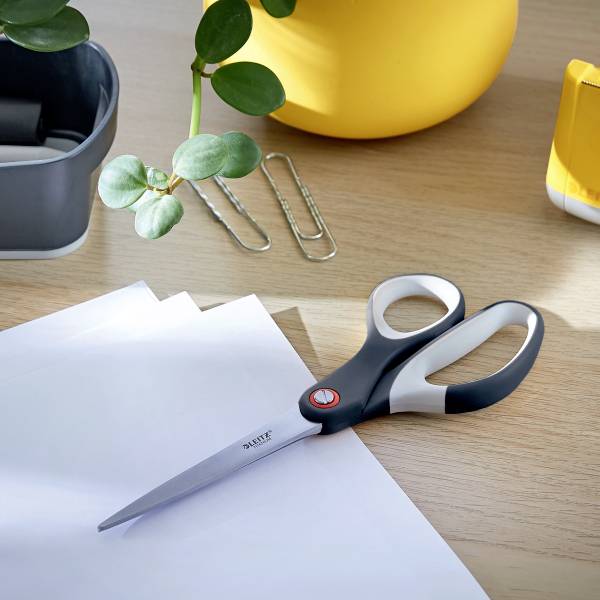 Eine Schere liegt auf einem Schreibtisch neben zwei Büroklammern, Pflanzenblättern, einem gelben Behälter und einem Blatt Papier.
