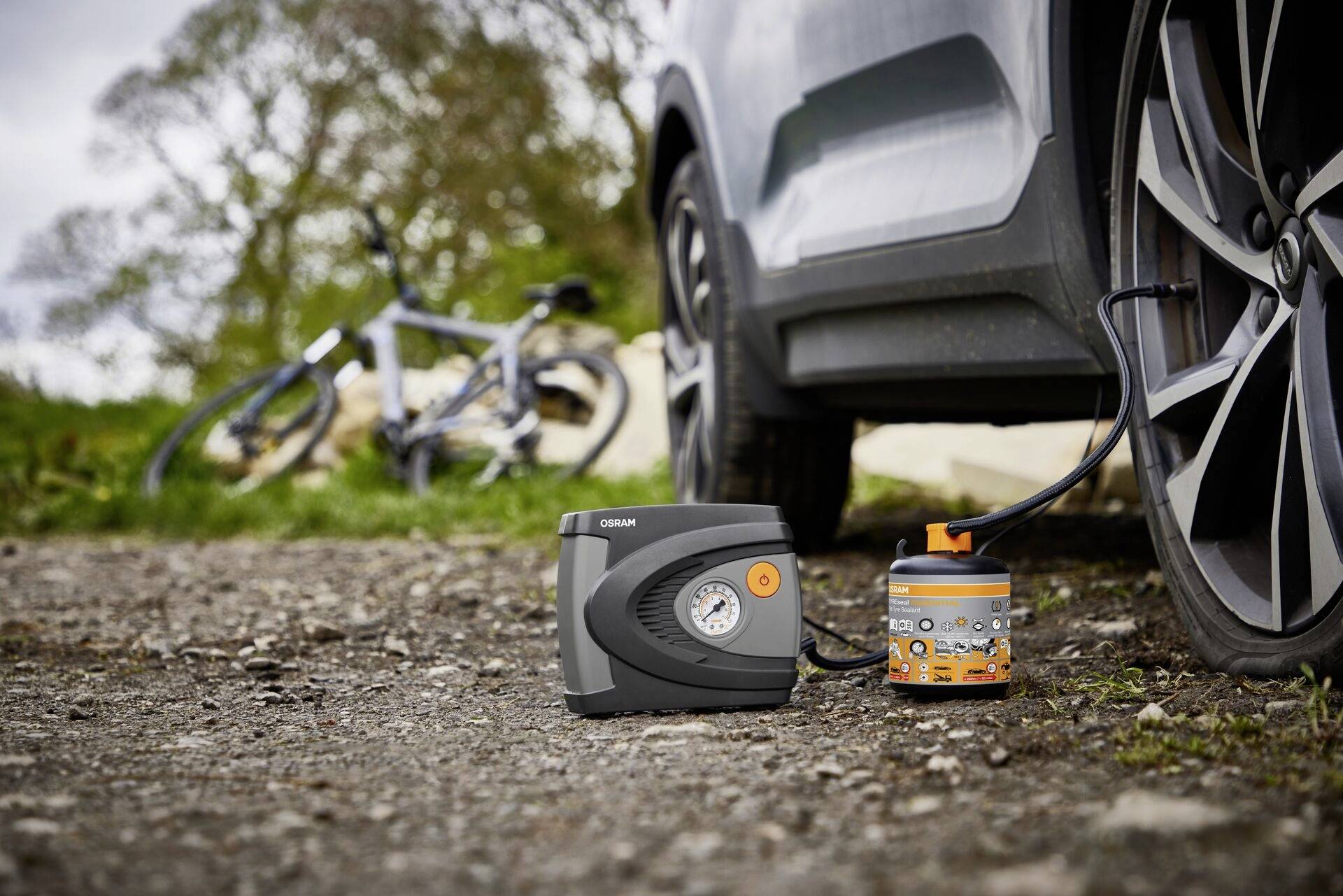 Ein Reifen eines Autos wird mit einem tragbaren Kompressor aufgepumpt, im Hintergrund ist ein Fahrrad auf einem Feldweg.