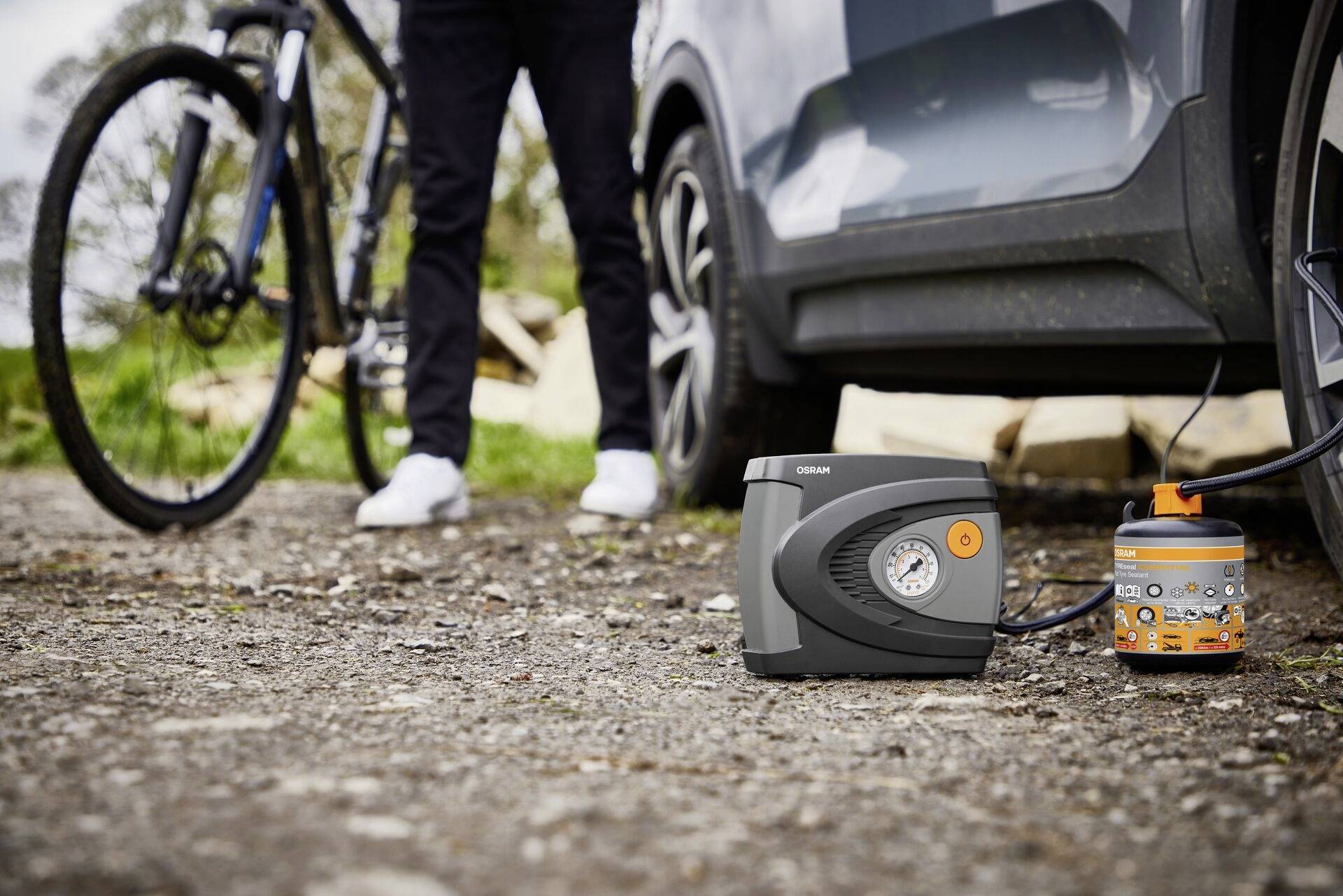 Eine Person steht neben einem Auto, während ein Reifen mit einem tragbaren Kompressor aufgepumpt wird. Ein Fahrrad lehnt daneben.