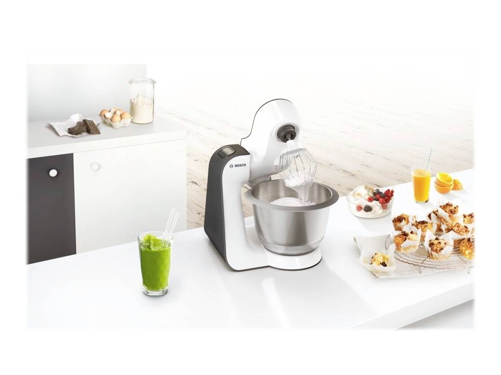 Küchenmaschine auf Tisch beim Schlagen von Teig. Daneben ein Glas mit grünem Smoothie, Muffins, Orangensaft und ein weiterer Drink.