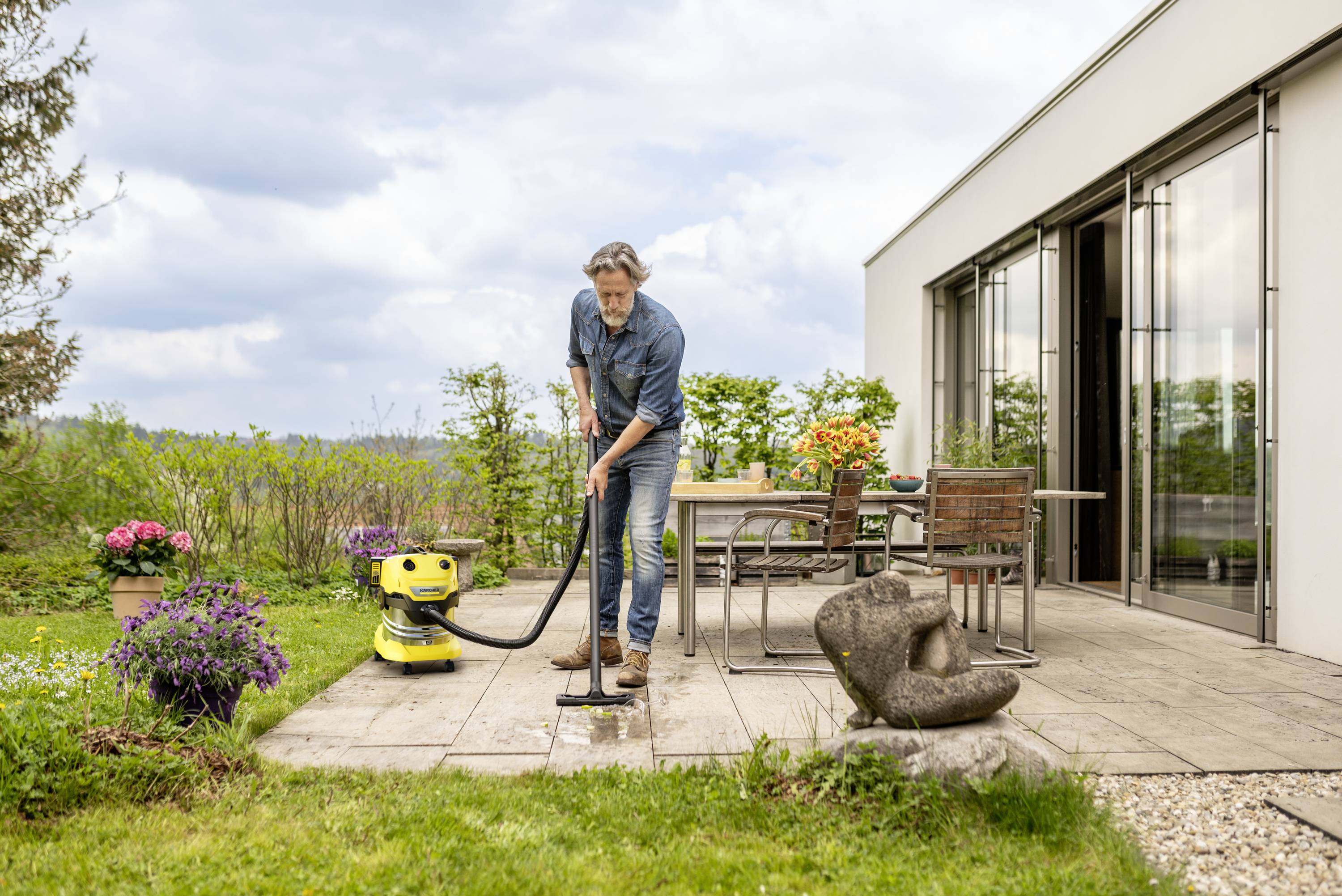 Ein Mann reinigt eine Terrasse mit einem Hochdruckreiniger, umgeben von Pflanzen und Gartenmöbeln. Der Himmel ist bewölkt.