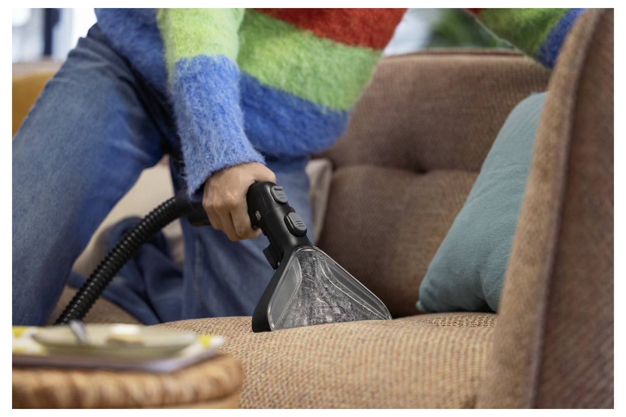 Eine Person in einem bunten Pullover benutzt einen Handstaubsauger auf einem braunen Sofa und veranschaulicht die Hausreinigung.