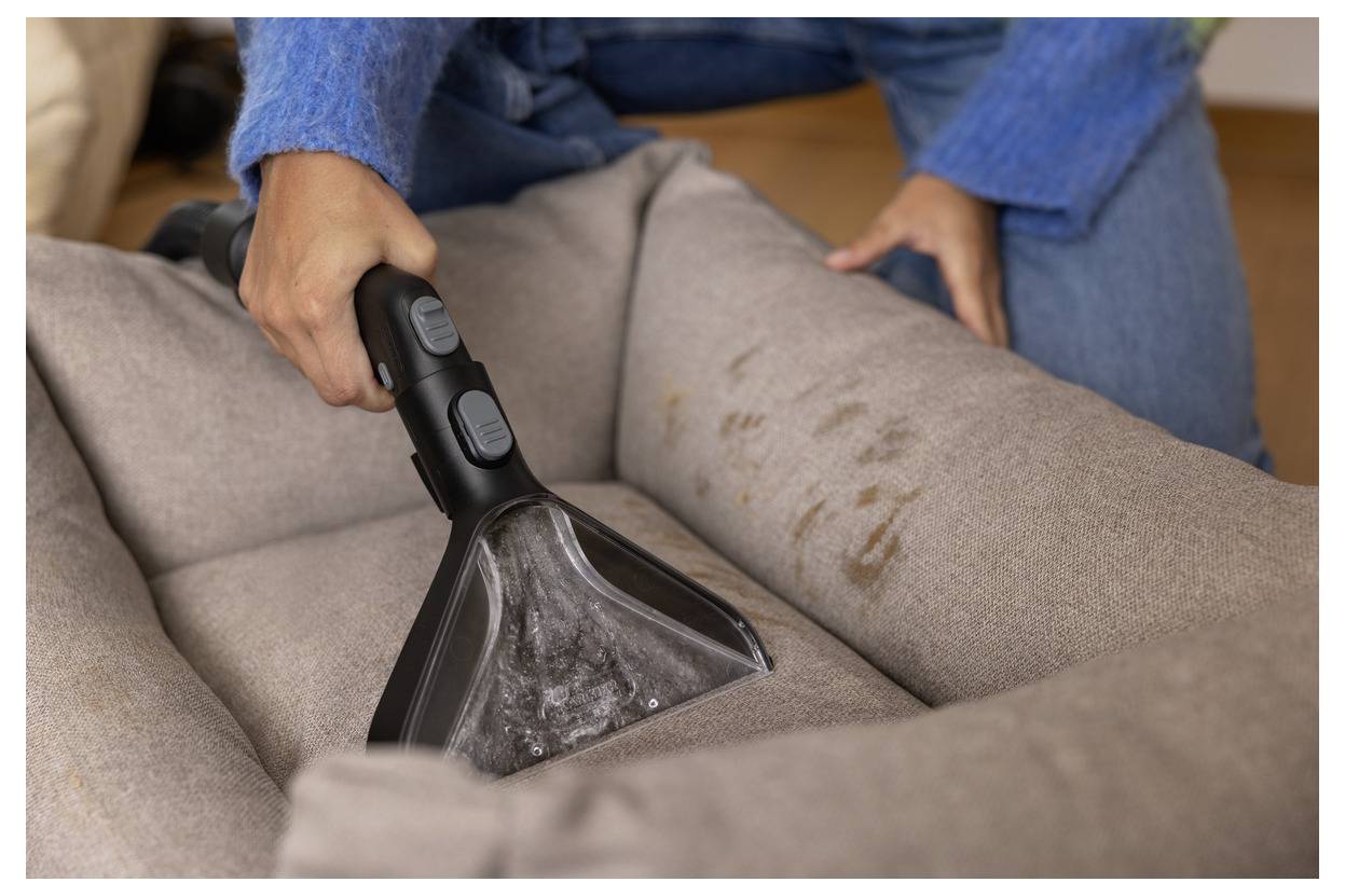 Eine Person benutzt einen tragbaren Staubsauger mit Bürstenaufsatz, um ein Haustierbett mit sichtbaren Flecken zu reinigen, und trägt dabei einen blauen Pullover und Jeans.