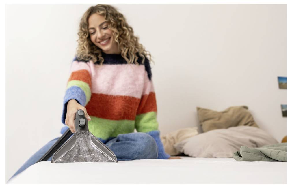 Eine Person mit einem bunten Pullover bedient einen Staubsauger auf einem weißen Bett und lächelt. Im Hintergrund sind Kissen zu sehen.