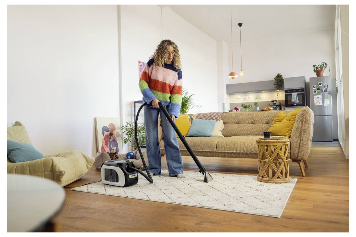 Eine Person staubsaugt ein gemütliches Wohnzimmer mit modernem Design, das eine beige Couch, bunte Kissen, einen gemusterten Teppich und eine stilvolle Lampe umfasst.