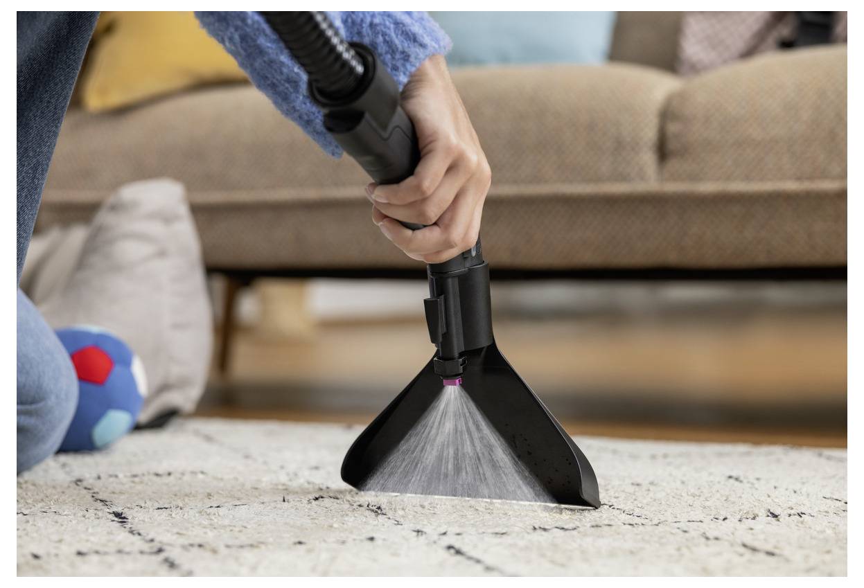 Eine Person saugt einen Teppich mit einem Handstaubsaugeraufsatz, wobei sie sich darauf konzentriert, Schmutz in der Nähe eines Sofas und eines bunten Balls zu entfernen.