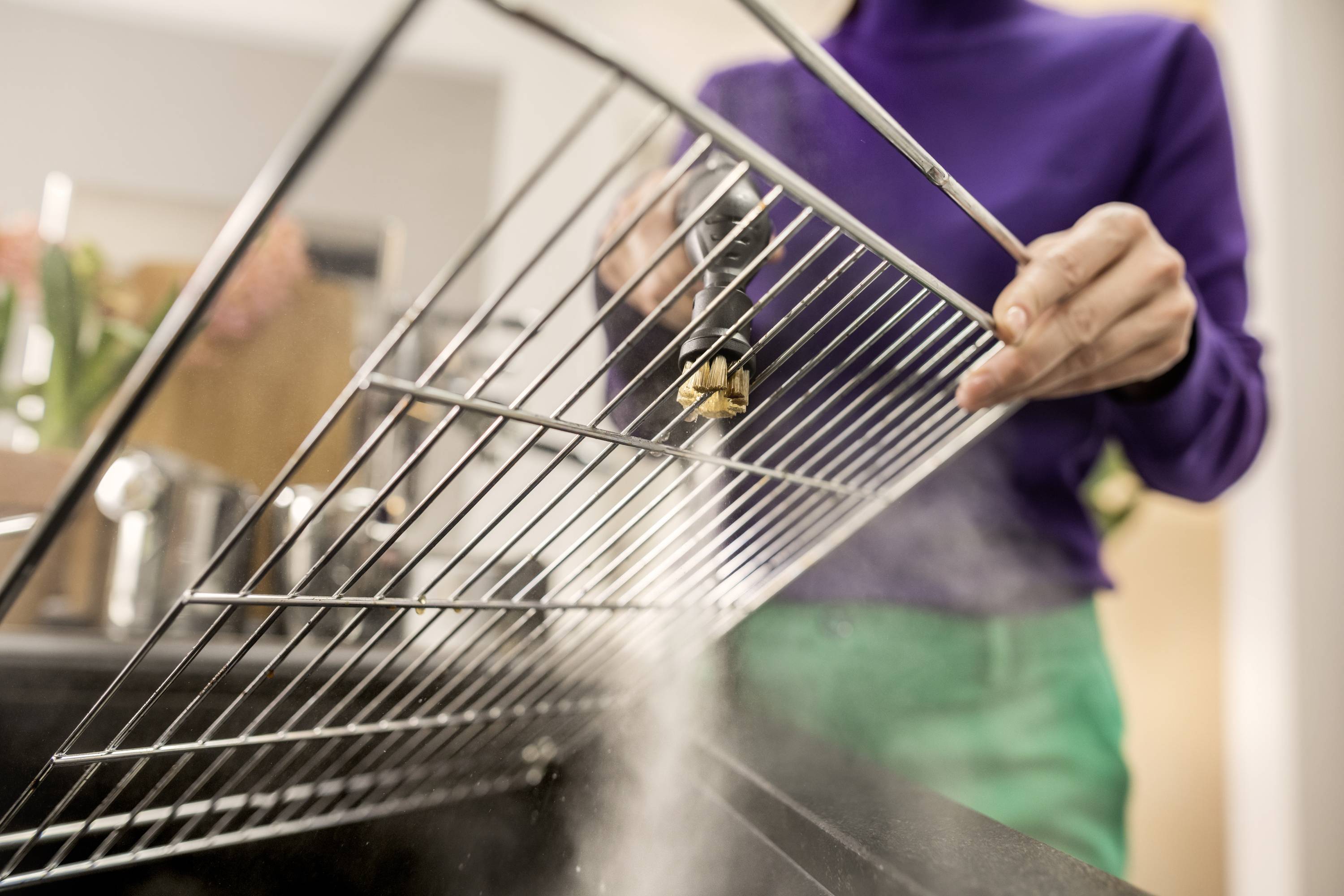 Eine Person reinigt ein Metallgitter über einem Waschbecken. Dampf steigt auf, während das Gitter mit Wasser abgespült wird.