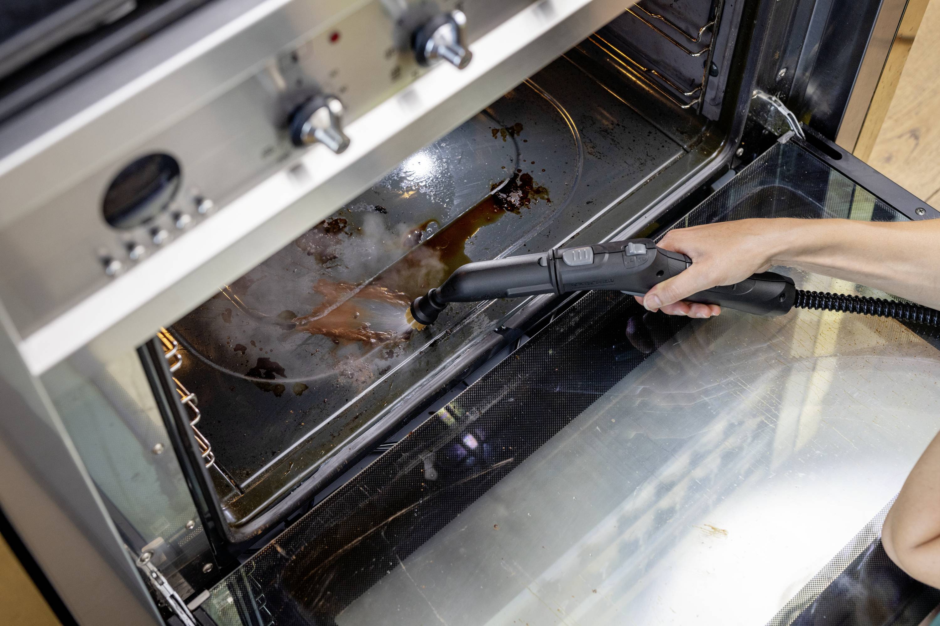 Eine Person reinigt mit einem Dampfreiniger einen schmutzigen Backofen. Verbrannte Essensreste auf dem Ofenboden werden entfernt.