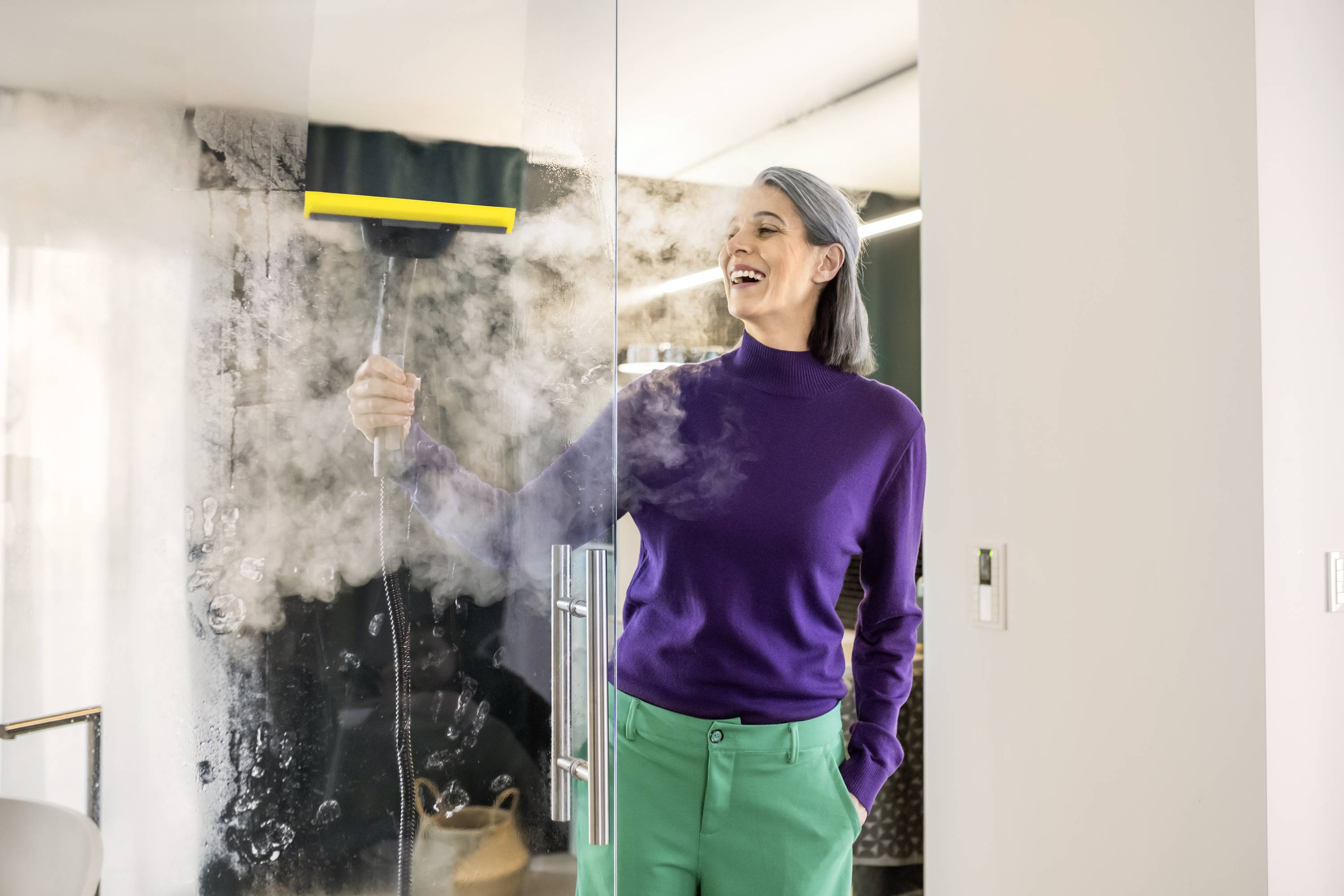 Eine lächelnde Person reinigt mit einem Dampfreiniger ein Glasfenster in einem hellen Raum.