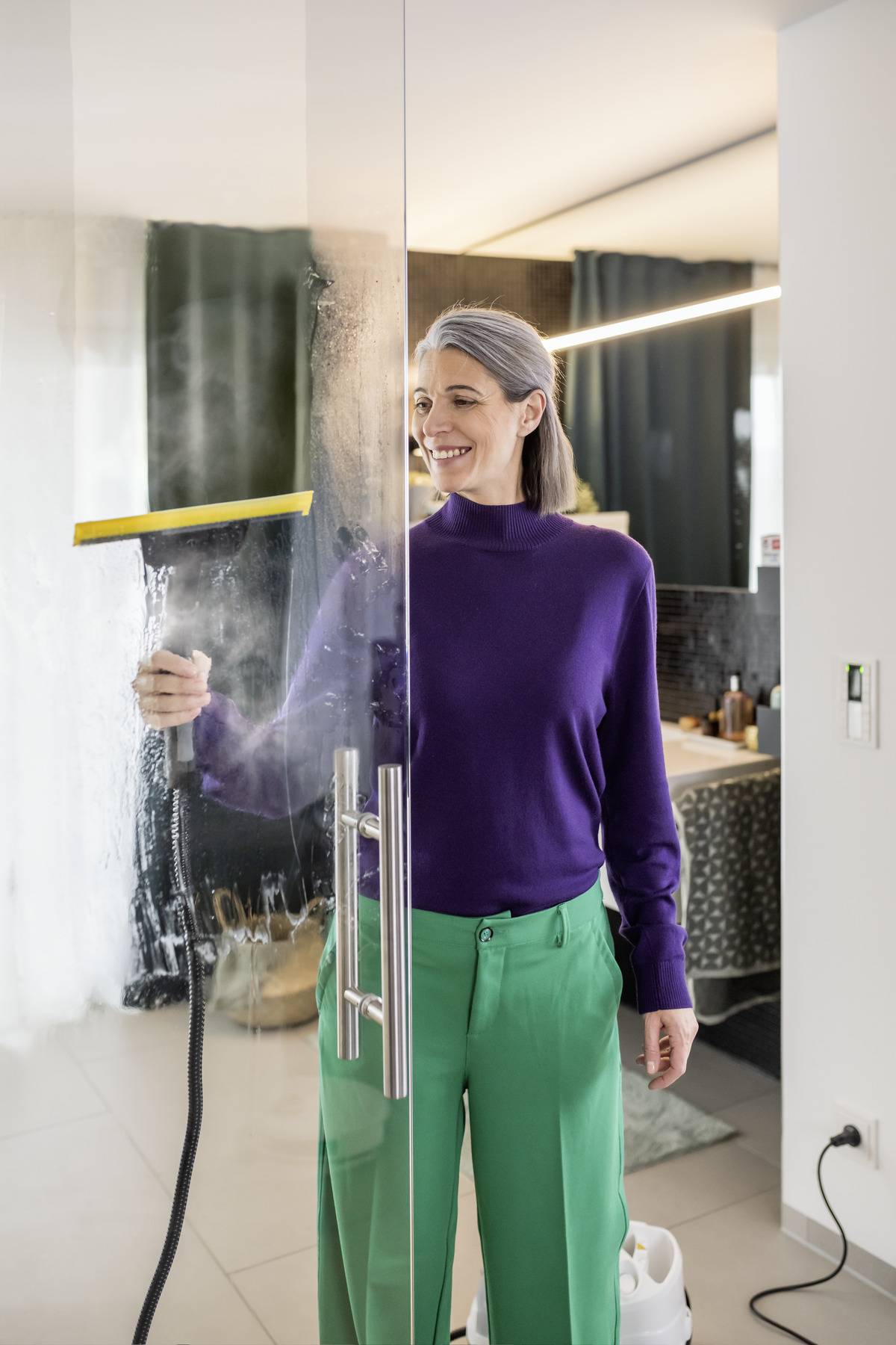 Eine Person, die eine Glastür mit einem Abzieher in einem modernen Zuhause reinigt. Sie lächelt und befindet sich offensichtlich in einem Badezimmer oder einer ähnlichen Umgebung.