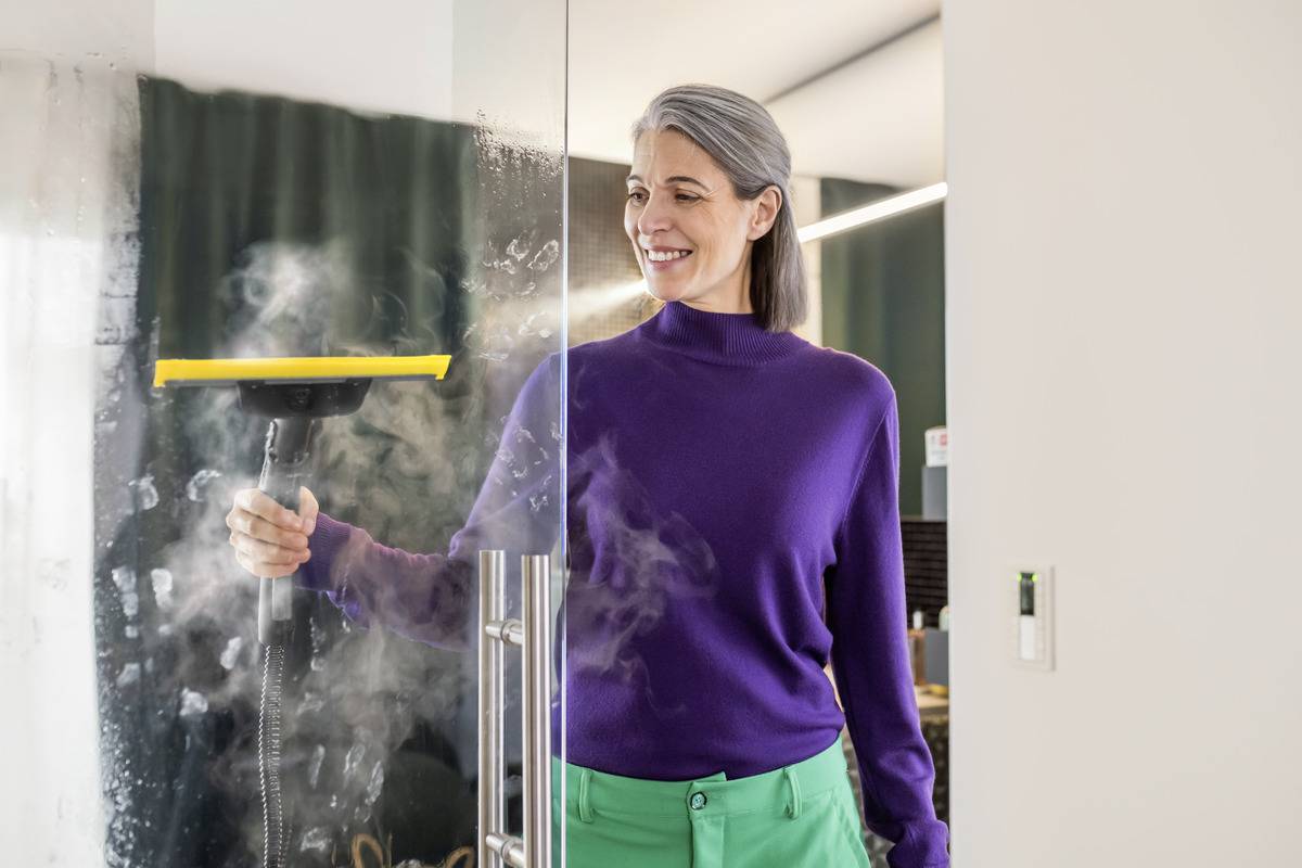 Eine Person mit grauen Haaren, die einen lila Pullover trägt, reinigt eine Glastür der Dusche mit einem Dampfreiniger. Dampf steigt auf, während sie arbeitet.