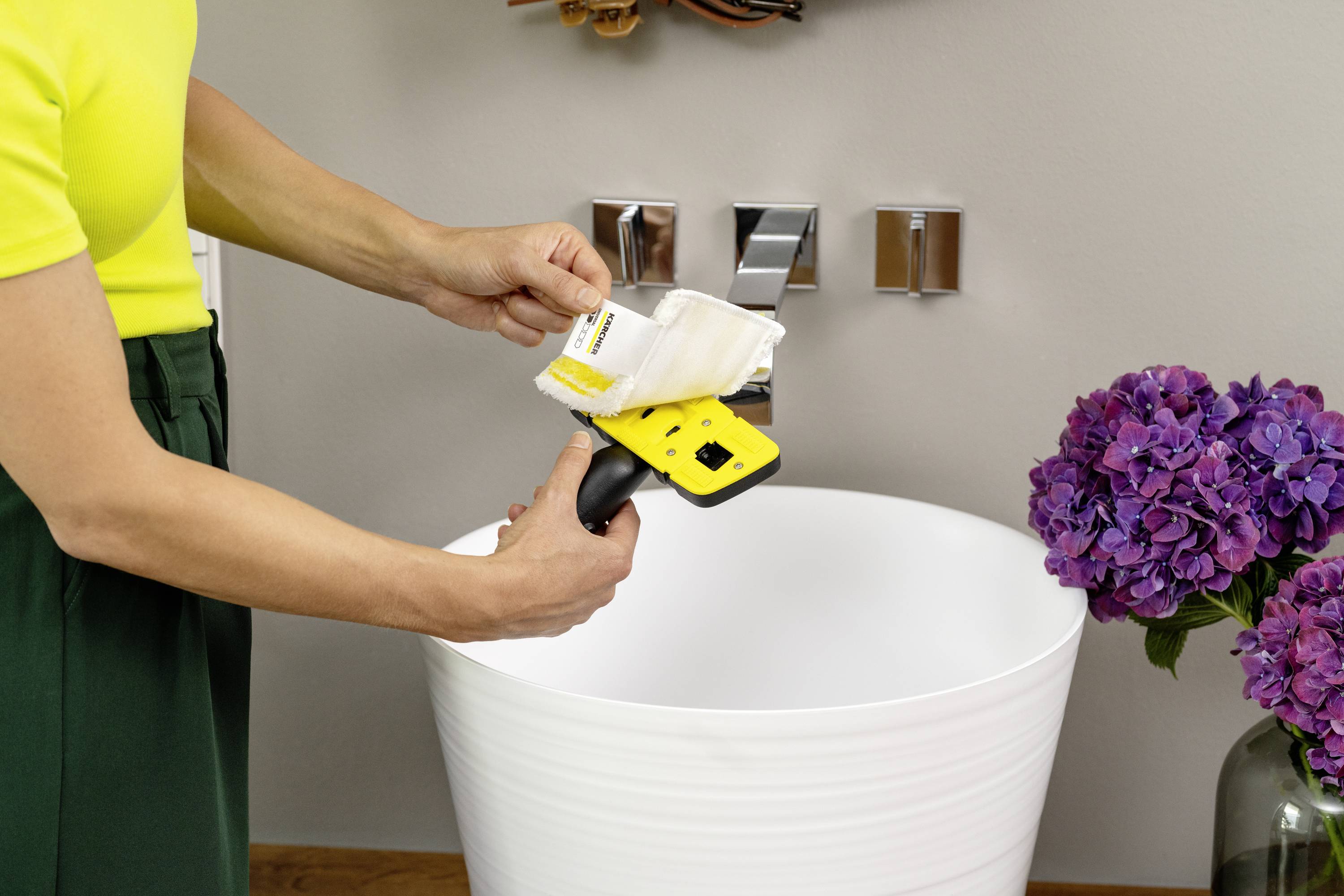 Eine Person reinigt einen gelben Fensterreiniger mit einem weißen Tuch über einem Waschbecken. Rechts im Bild steht ein Blumenstrauß in einer Vase.
