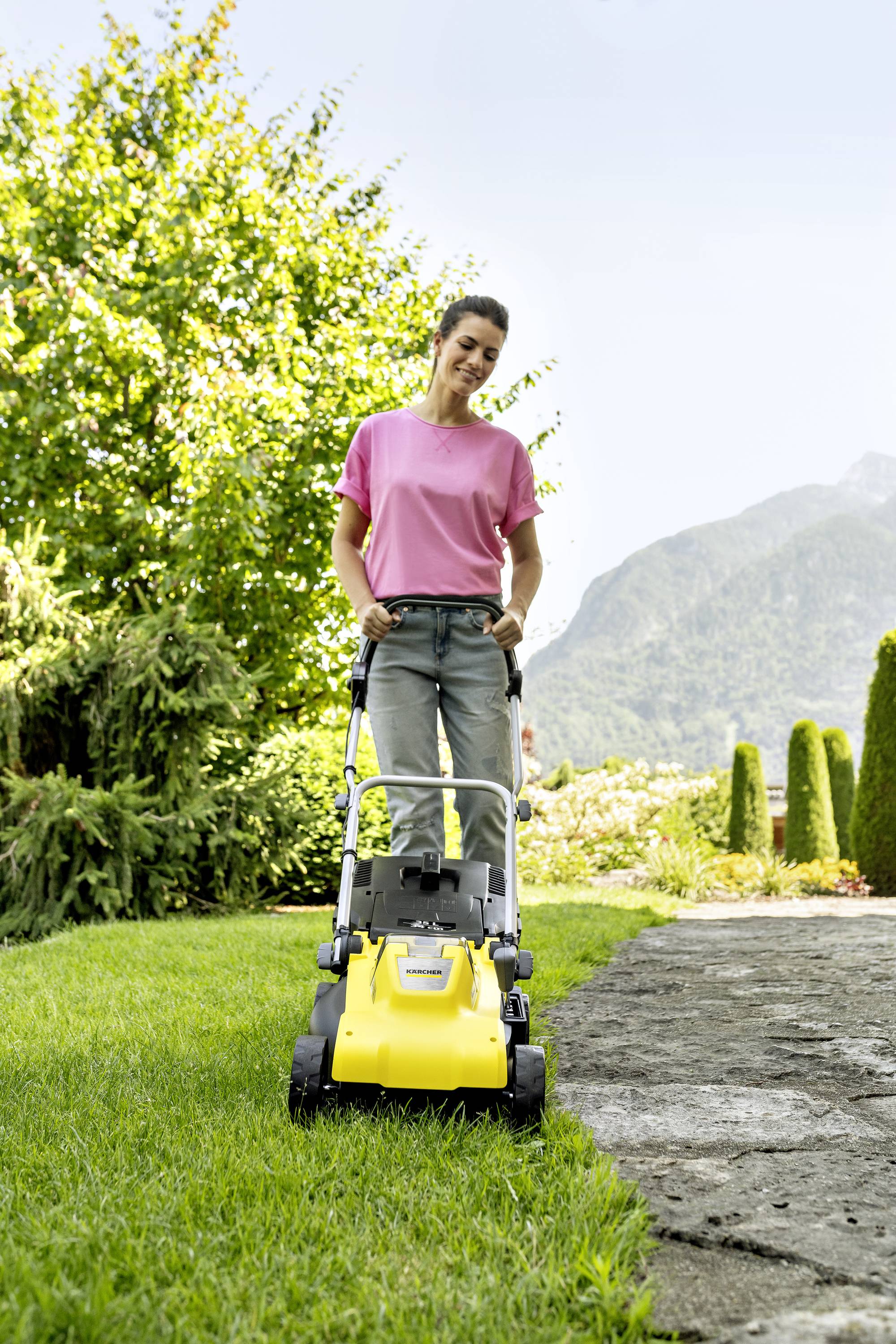 Eine Frau mäht den Rasen mit einem gelben Elektro-Rasenmäher in einem Garten, umgeben von Bäumen und Bergen im Hintergrund.