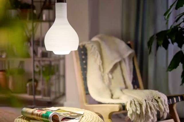 Eine gemütlich eingerichtete Ecke mit einem hellen Sessel, bedeckt mit einer Decke, steht neben einem kleinen Tisch mit Zeitschriften. Eine Lampe leuchtet von oben herab.