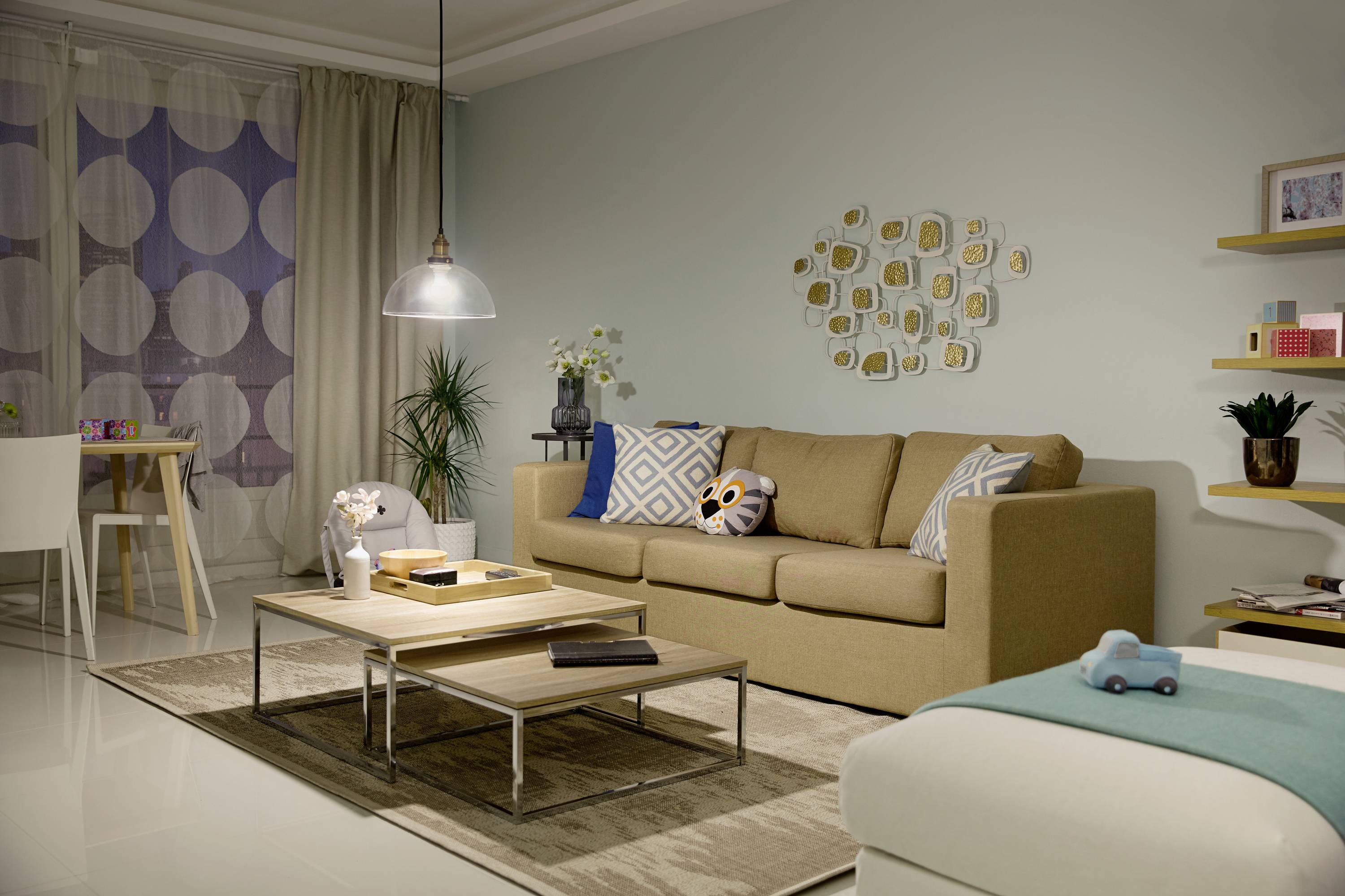 Ein modernes Wohnzimmer mit beigem Sofa, Couchtisch, Wanddeko und Pflanzen. Im Hintergrund ein Esstisch und eine Lampe.