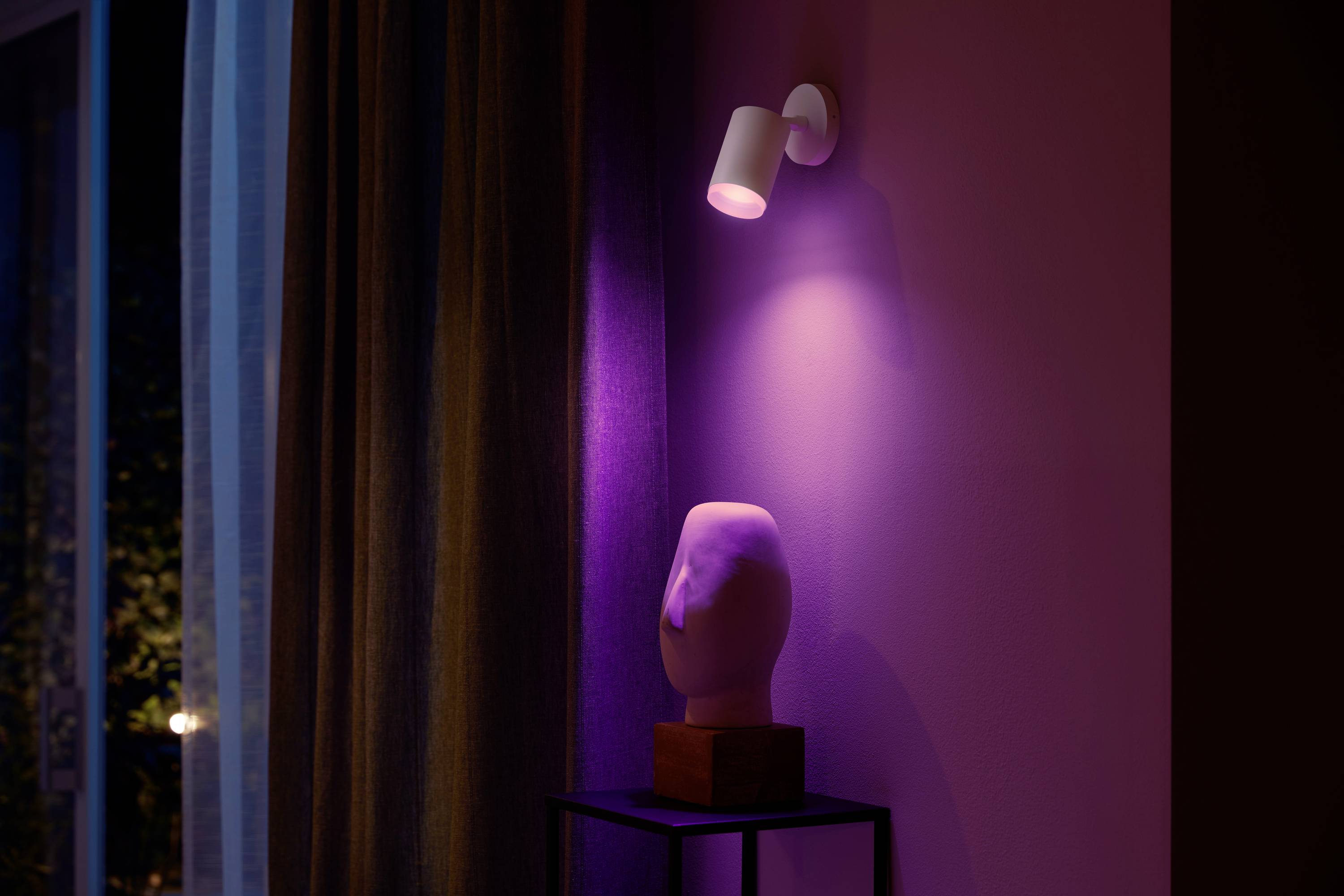 Ein weißer Scheinwerfer beleuchtet eine abstrakte Skulptur auf einem Tisch in einem Raum mit violettem Licht und Vorhang im Hintergrund.