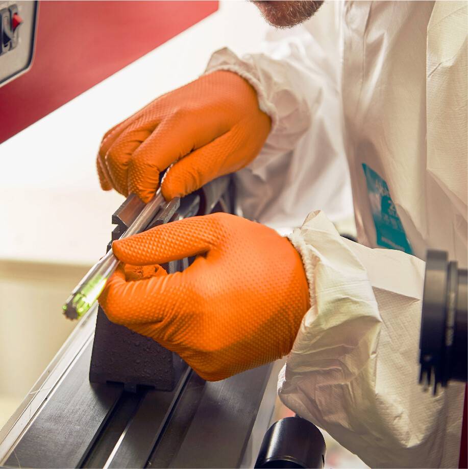 Eine Person in Schutzkleidung und orangefarbenen Handschuhen bearbeitet ein metallisches Bauteil mit einem Werkzeug in einer Werkstatt.