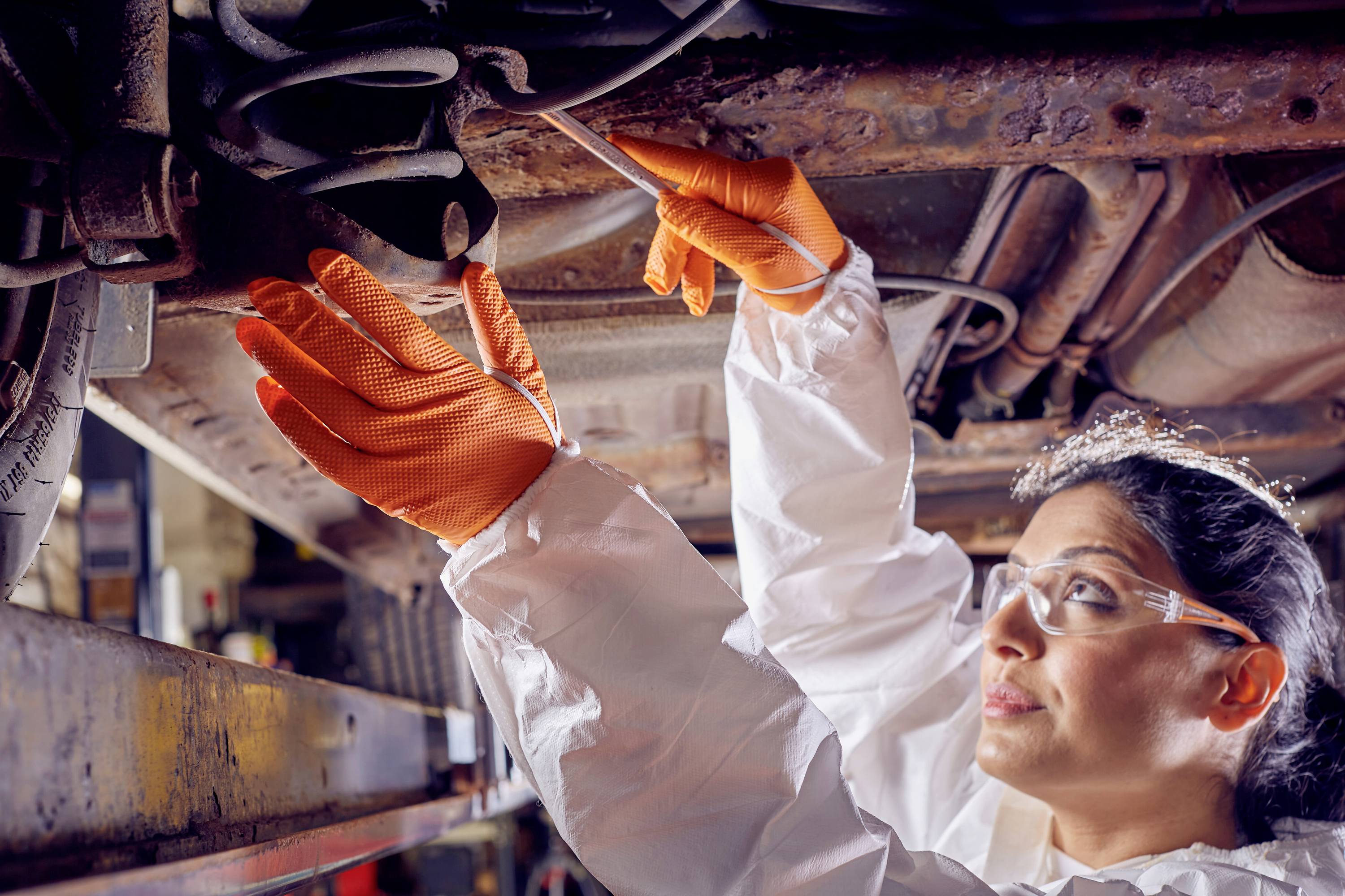 Eine Mechanikerin in Schutzkleidung und Handschuhen inspiziert die Unterseite eines Fahrzeugs in einer Autowerkstatt.